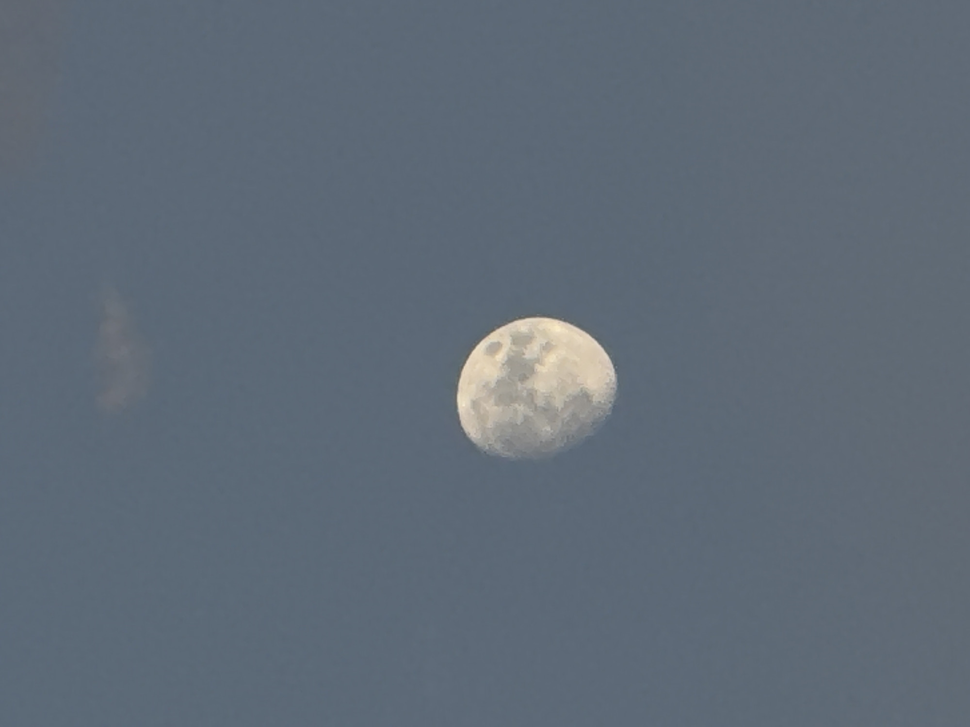 A waxing gibbous moon is visible in a hazy blue sky. The moon is off-center, towards the bottom right of the frame, and displays visible craters and surface features. A faint, wispy cloud is present in the upper portion of the sky, directly above the moon. The overall scene suggests an outdoor environment, likely at dusk or dawn, given the color of the sky. There are no people, structures, or any other discernable objects or elements within the frame. The image is a close-up shot focusing on the moon and the sky.