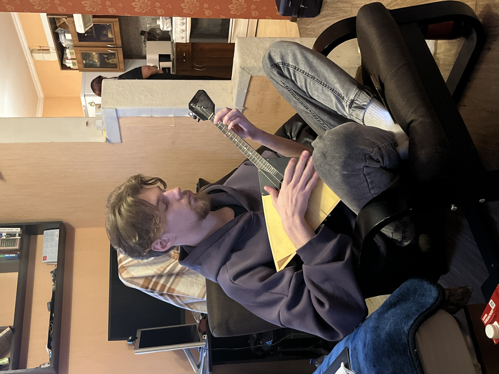 A man with light brown hair and a beard is sitting in a chair, playing a triangular stringed instrument resembling a balalaika. He is wearing a purple hoodie and gray jeans, with white socks visible. The chair is upholstered in brown fabric. The background shows a living room setting with a television, a bookshelf with books, and a kitchen area in the distance. The kitchen has cabinets and appliances. Another person is partially visible in the kitchen.
There's no specific evidence that this image was taken in Rostov-on-Don, Russia.