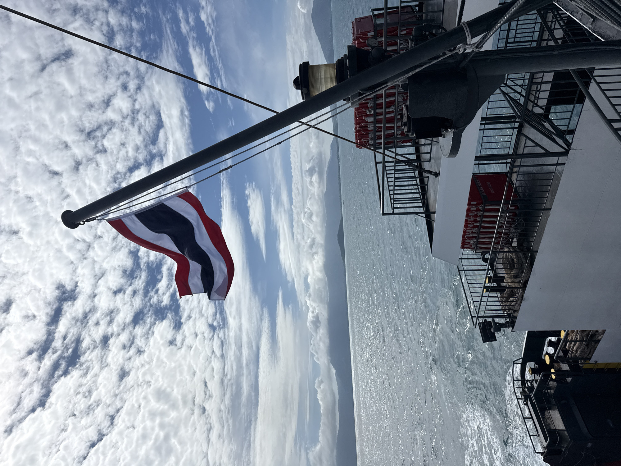 The image captures a view from a ferry in Thailand. The Thai flag is prominently displayed on a flagpole, waving in the wind against a backdrop of a partly cloudy sky. The ferry itself is visible, with railings and safety equipment, including life preservers, lining the deck. The sea is a tranquil blue, with distant islands or a coastline visible on the horizon, shrouded in mist or clouds. The overall scene suggests a peaceful journey across the water.