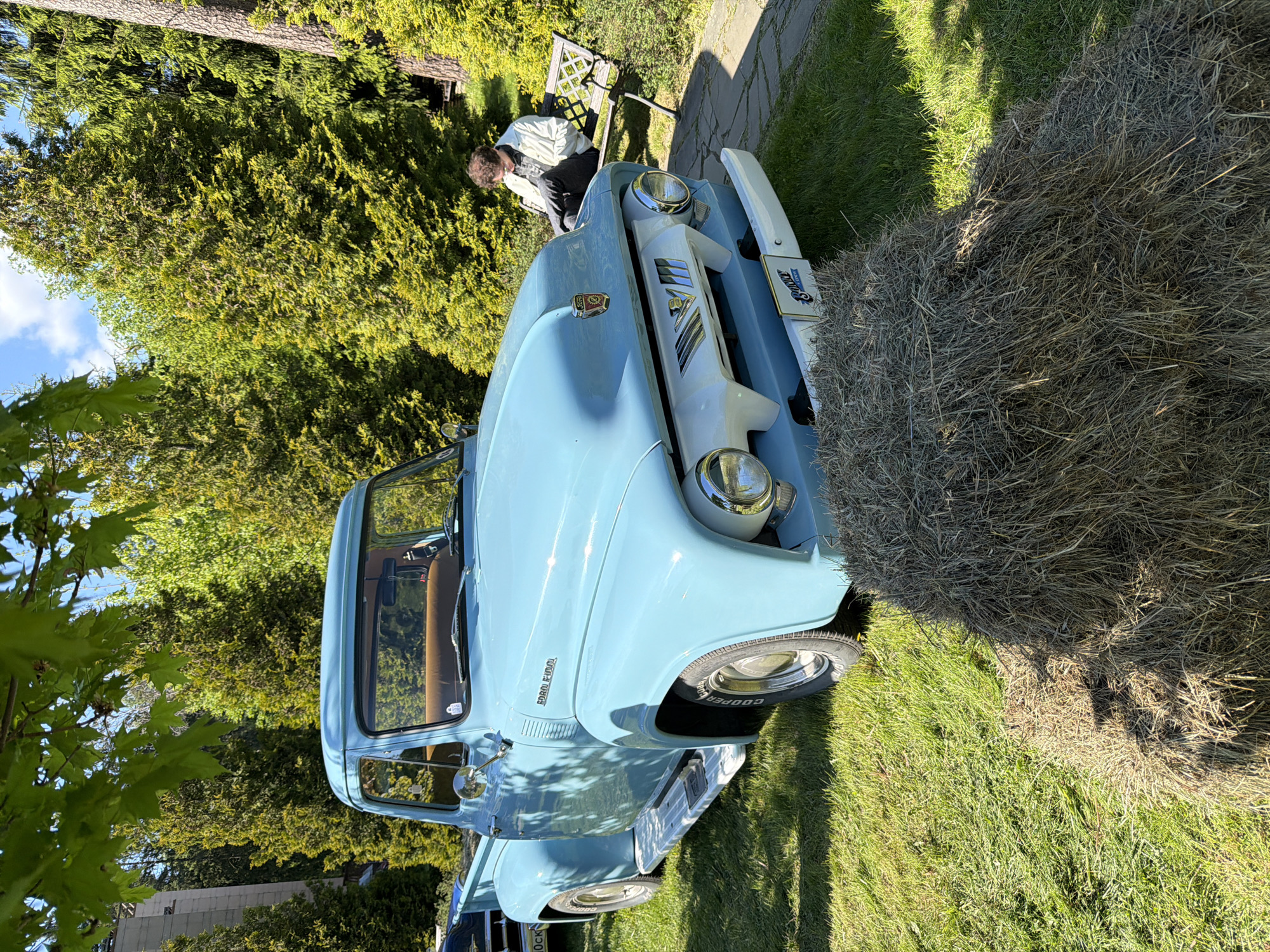 A light-blue vintage Ford F-100 pickup truck is parked on a patch of green grass beside a bale of hay in the city of Repino, Russia. A person is sitting on a bench in the background. The area is shaded by trees.
