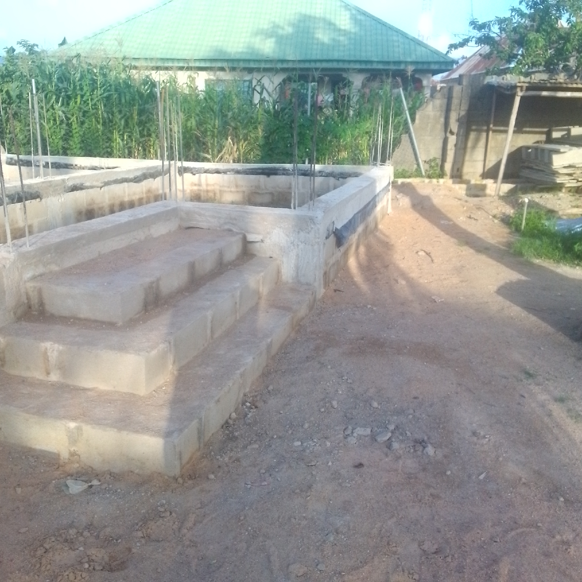 The image depicts an outdoor construction scene in Jalingo, Nigeria. The foreground features the base of a structure, likely a building, with concrete walls and rebar protruding. A set of concrete steps leads up to this base. The construction appears to be in an early stage, with loose soil and gravel covering the ground.
In the background, a building with a green, corrugated metal roof is visible, partially obscured by tall green vegetation, possibly corn stalks. To the right, a separate, open-sided structure with a similar metal roof stands. Several stacks of wooden planks are piled near this structure. The sky is overcast, suggesting either early morning or late afternoon, or possibly a cloudy day. The overall setting is rural or semi-rural, indicative of ongoing development. There is no visible text in the image.