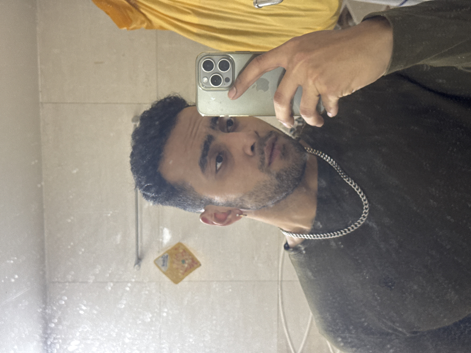 The image shows a man with dark hair and a beard taking a selfie in a bathroom mirror. He is wearing a dark green shirt and a silver chain necklace. He is holding an iPhone in his hand, and his reflection is visible in the mirror, which also shows a tiled wall, a yellow shirt hanging above, a small framed picture, and a blurry view of the bathroom. There is no information available in the image to determine if the photograph was taken in the city of Kolkata, India.