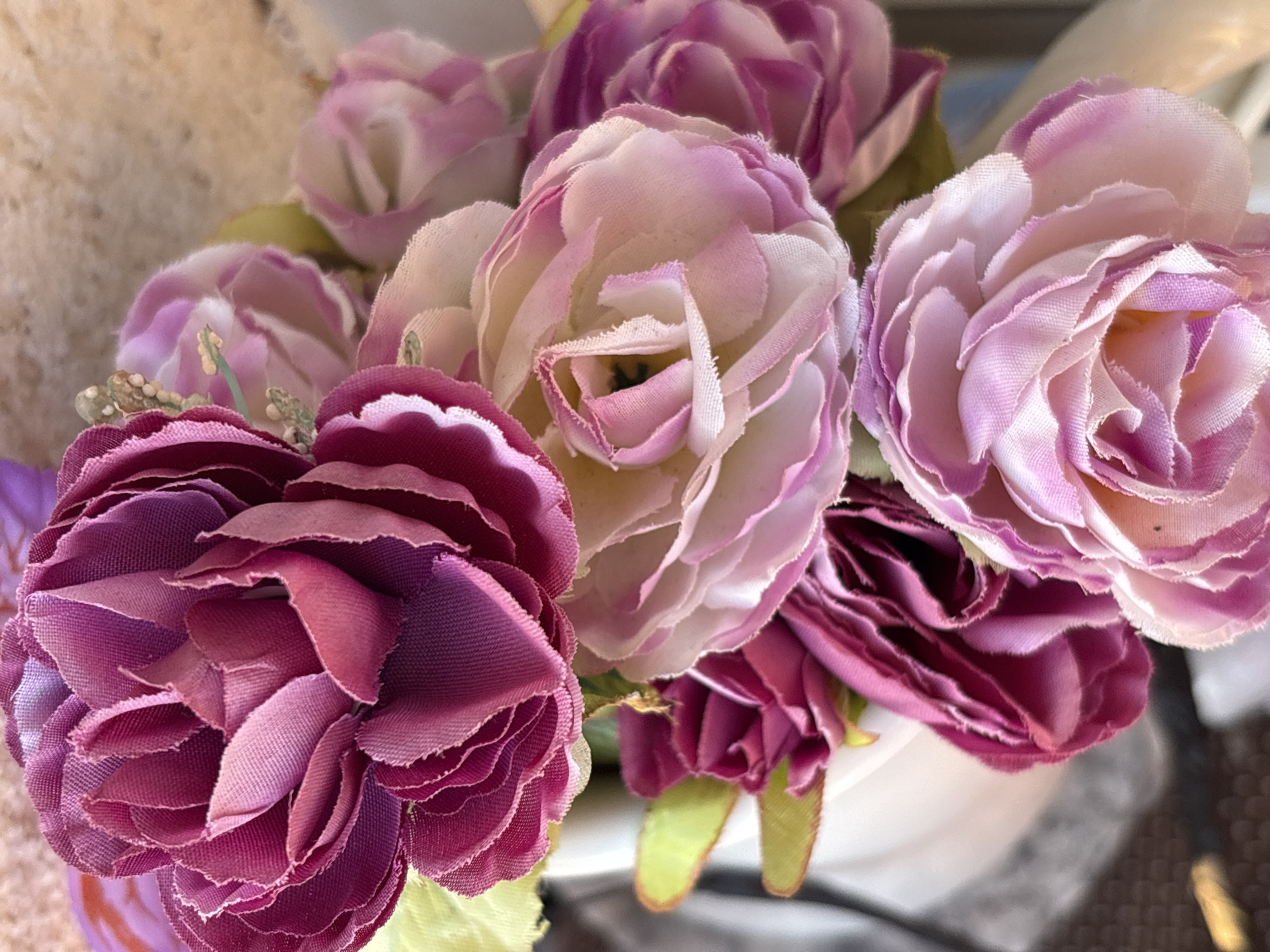 The image shows a close-up view of a bouquet of artificial flowers in shades of pink and purple. The flowers have multiple layers of fabric petals, with a textured, slightly frayed edge. Some of the flowers are fully bloomed, while others appear to be in bud form. The bouquet is arranged in a way that suggests it might be a decorative item, possibly in a home or a shop. The background is blurred, but it appears to be an indoor setting with some natural light. There are no people or discernible actions in the image. The image does not provide any specific visual cues that would place it in Osterode am Harz, Germany, or any other specific location.
