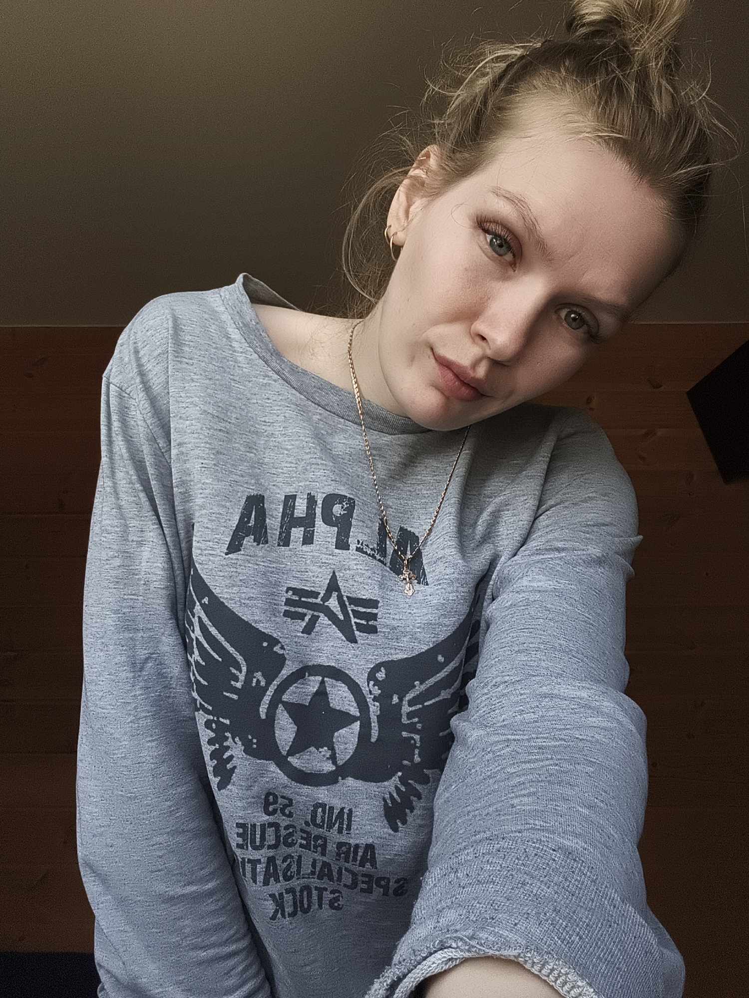 A woman is depicted indoors, wearing a grey long-sleeved shirt with a graphic design featuring the word "ALPHA," an emblem with wings and a star, and reversed text reading "2LOCK SPECIALIST AIR RESCUE IND 2Q." She has blonde hair styled in a messy bun and is wearing a gold necklace and earrings. The background consists of a dark brown wooden wall and ceiling. The scene is dimly lit. The location is not discernible from the image.