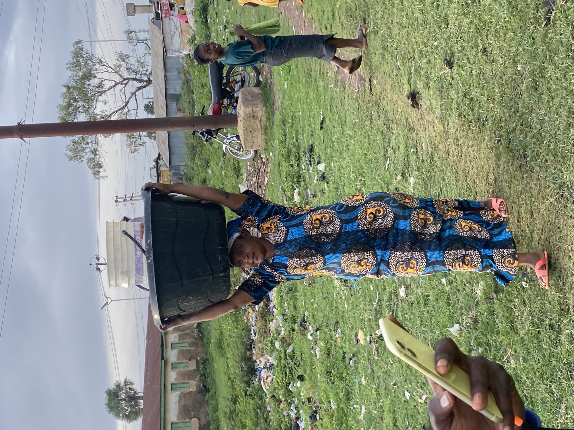 A woman walks across a grassy area in Bukuru, Nigeria, carrying a large black tub on her head. She is wearing a blue and orange patterned dress and pink sandals. To her right, a young girl in a green shirt and dark skirt smiles at the camera, holding a green bag. Behind them, a red motorcycle is parked next to a utility pole. The ground is littered with trash, suggesting an urban environment. In the foreground, a hand holds up a yellow smartphone. The sky is overcast.