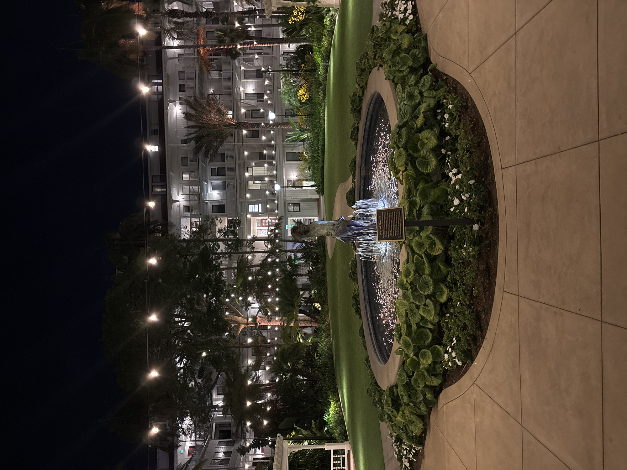 At night, a long rectangular building with multiple floors and evenly spaced illuminated windows stands beside a pathway leading to a fountain in Coronado, United States of America. The building appears to be a hotel or residential complex. Palm trees are positioned between the building and the pathway, creating a layered visual field. Overhead string lights are suspended from the trees, enhancing the ambient lighting. A round fountain, bordered by lush greenery, sits to the right of the pathway. A signpost is placed near the fountain. The scene is set in Coronado, United States of America.