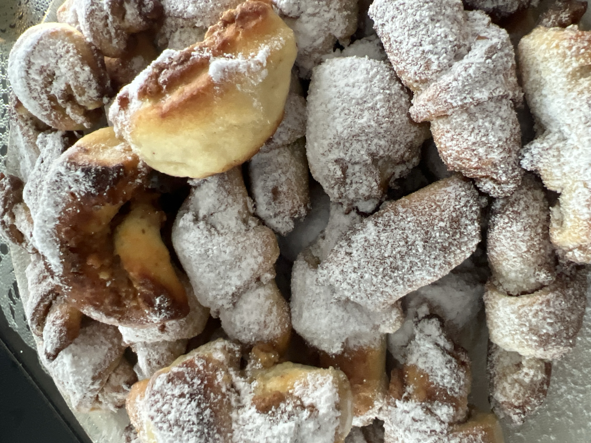 A close-up, overhead view showcases a pile of freshly baked crescent-shaped pastries dusted generously with powdered sugar. The pastries, varying in shade from golden brown to a lighter dough color, exhibit a slightly irregular, homemade appearance. Some have a noticeable shine, hinting at a glaze or butter content. The powdered sugar forms a fine, white coating, obscuring some of the finer details of the pastry's texture. The background is dark, suggesting a surface like a table or a baking tray. This scene is likely from Dillingen an der Donau, Germany, a locale known for its traditional bakeries and pastry shops.