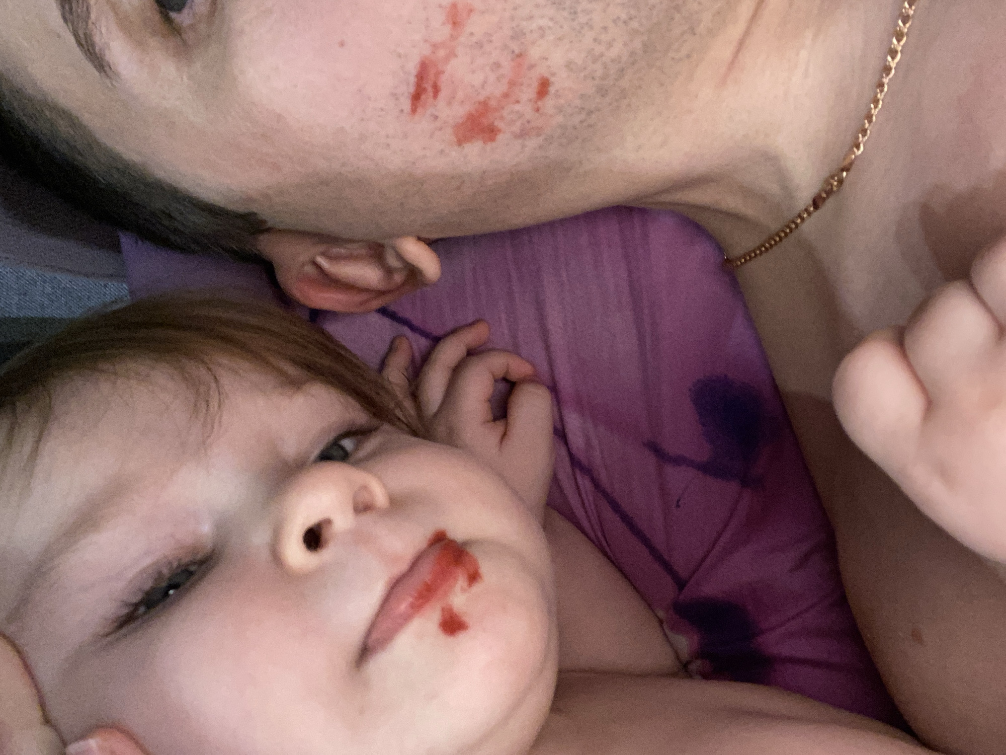 The image shows a close-up of a man and a baby lying down. The man has red stains on his face, and the baby has red stains around its mouth. The background is a purple surface with dark stains. The location is unknown.