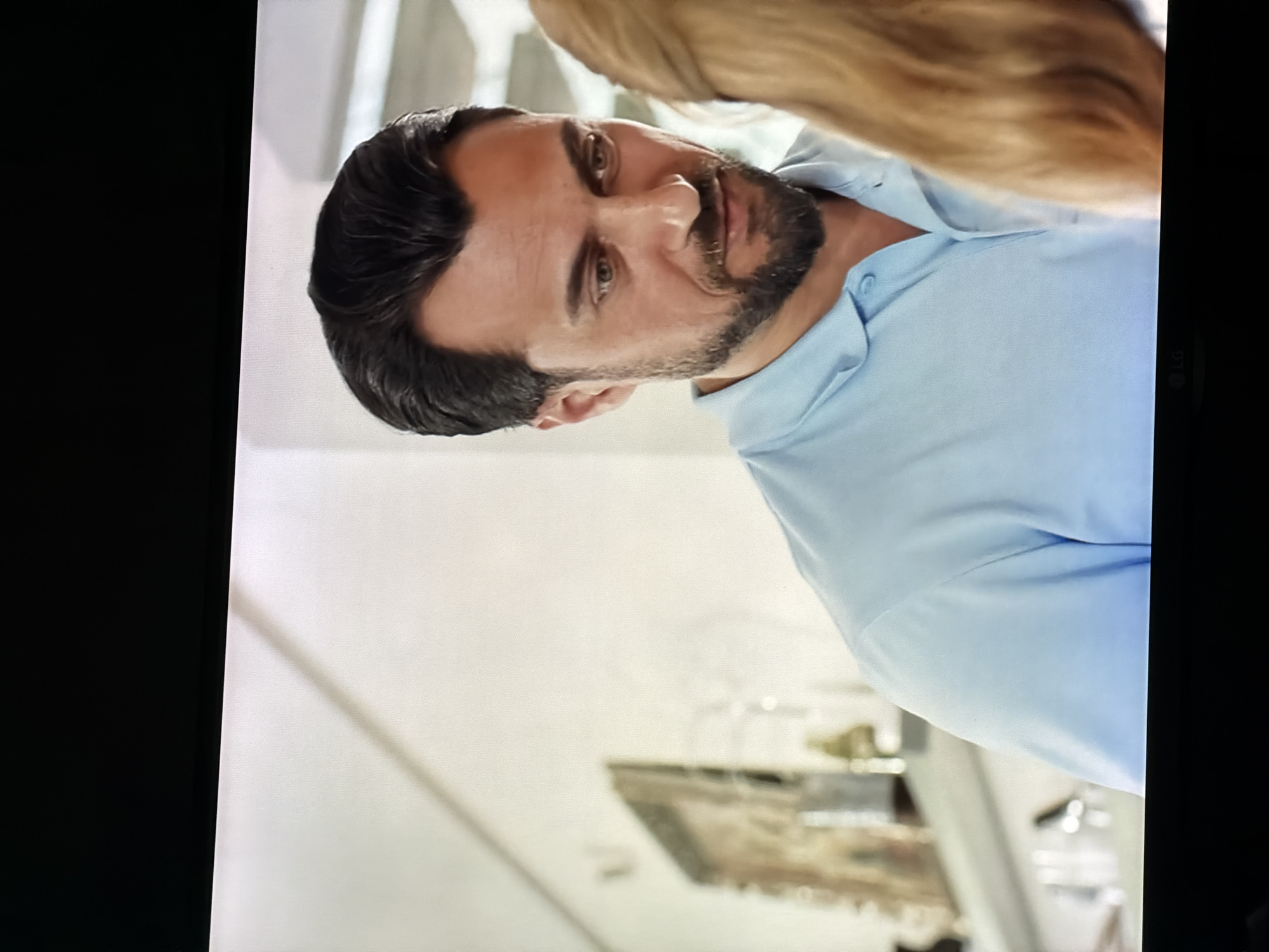 The image shows a medium shot of a man with dark hair and a beard, wearing a light blue polo shirt. He is looking down and to his right, with his head slightly tilted. His expression appears contemplative or concerned. To the right of the man, a portion of a blonde woman's hair is visible, suggesting they are interacting. The background is blurred, but appears to be an indoor setting with light-colored walls and shelves with decorative items. The man's gaze is directed towards something or someone out of frame. The lighting suggests a bright, indoor environment. There is no visible indication of the city of Semiluki, Russia in the image.