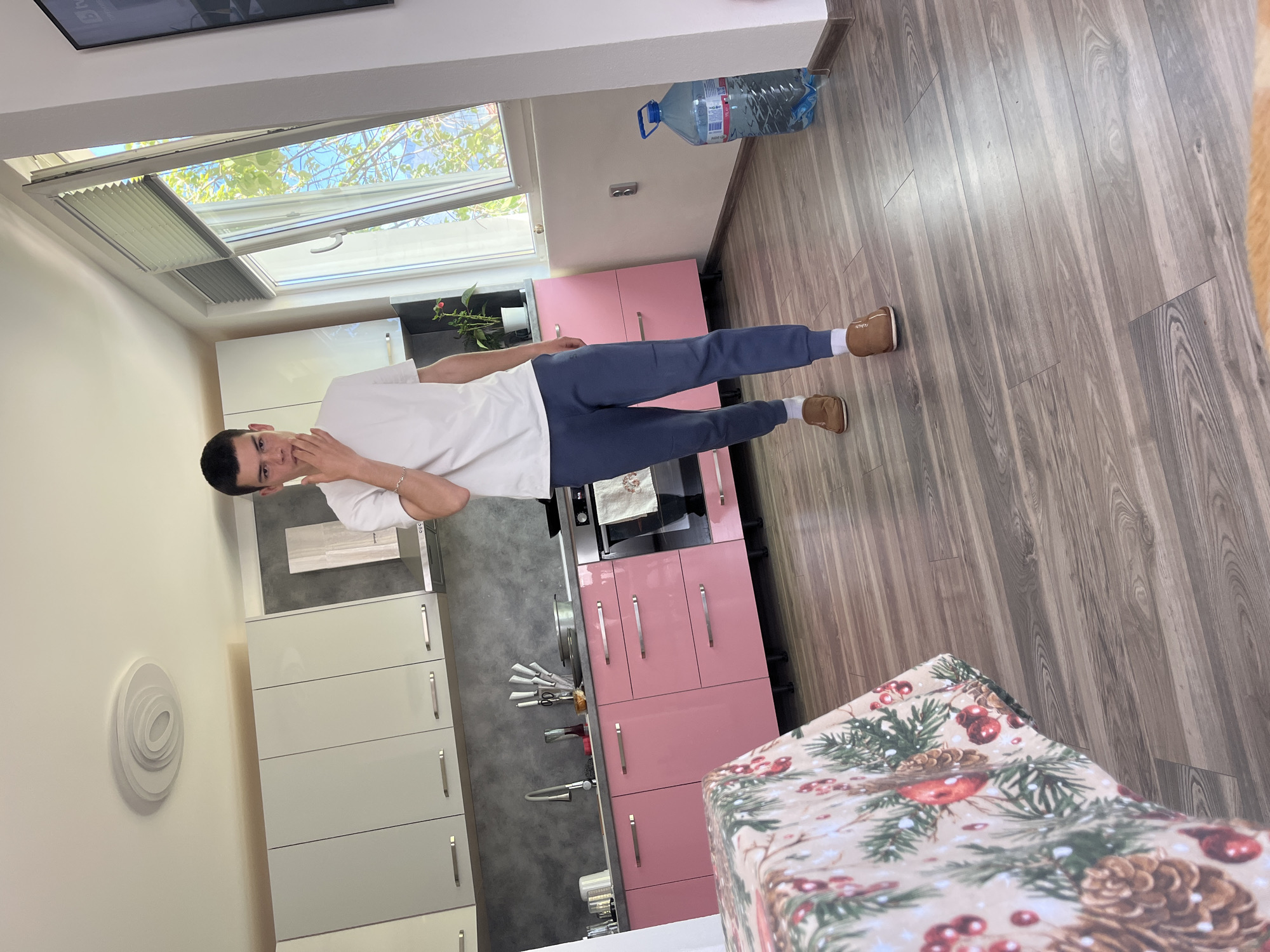 A man is standing in a kitchen. The kitchen is equipped with white and pink cabinets, a sink, an oven, and various kitchen utensils. A large bottle of water is placed on the wall. The flooring is light brown wood. A wrapped gift with a Christmas-themed pattern is positioned on a table. The location cannot be determined from the given information beyond it being a possible interior of a building, and therefore cannot be confirmed as being in Karavelovo, Bulgaria. The man is wearing a white shirt, blue pants, and brown shoes. He has his hand near his face.