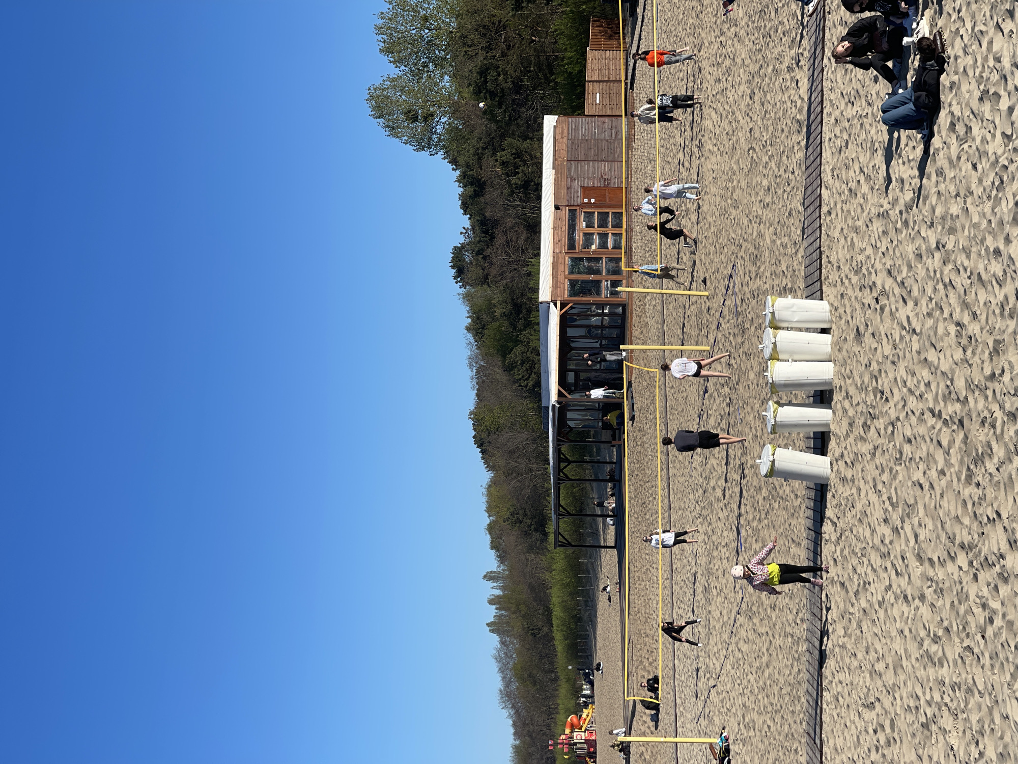 The image depicts a beach volleyball area in Gdańsk, Poland, with multiple courts and a small wooden building serving as a concession stand or clubhouse. The courts are occupied by people playing volleyball, while others are lounging or walking along the beach. The environment includes sandy beach, vegetation in the background, and a clear blue sky. There are volleyball nets and boundary lines marking the playing areas. The location appears to be a public beach with recreational facilities.