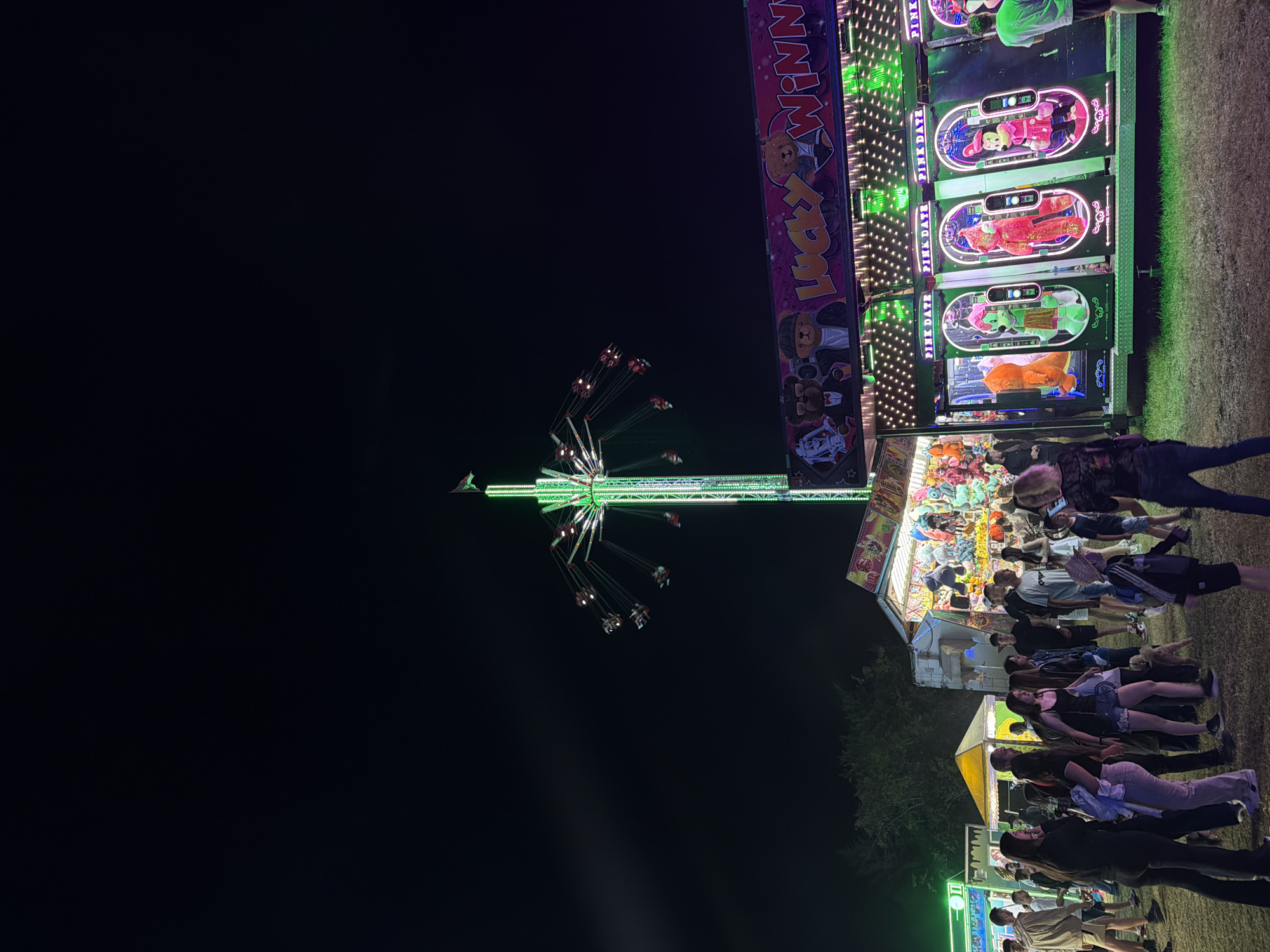 The image depicts an outdoor amusement park at night, likely in Sofia, Bulgaria, as indicated by the presence of a "Lucky Winner" game booth and other carnival attractions. A tall, illuminated swing ride, predominantly green, dominates the center of the frame, with several people visible on the spinning seats. To the right, a brightly lit game booth, also adorned with lights, features several plush toys inside glass enclosures, possibly for a claw machine game. The "Lucky Winner" sign is prominently displayed across the top of this booth, featuring cartoonish bear figures and the words "Lucky Winner" in a playful font.
In the foreground, a crowd of people are milling around, some dressed casually, suggesting a relaxed evening at the fair. The ground is covered in grass, and a few trees are visible in the background, indicating an outdoor setting. The overall atmosphere is one of lively entertainment, with the vibrant lights of the rides and games contrasting against the dark night sky. The image captures the essence of a fun-filled evening at a carnival, with a focus on the rides and games that are characteristic of such events.