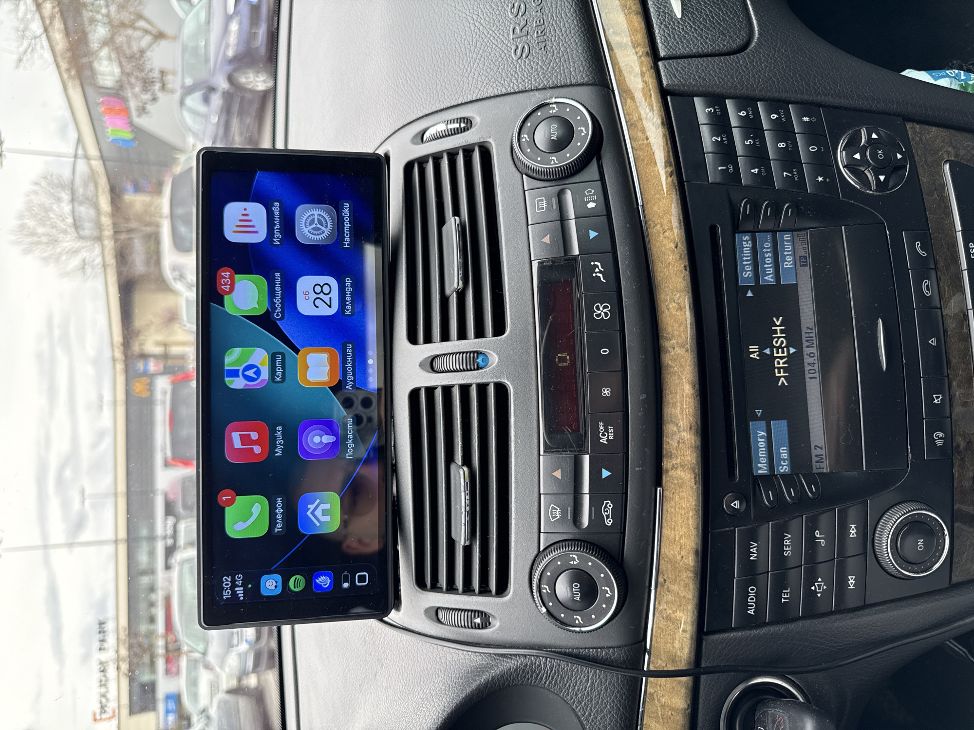 The image displays the interior of a vehicle, viewed from the driver's perspective, focusing on the dashboard and the scene outside the windshield. A large, rectangular aftermarket infotainment screen is centrally mounted above the car's existing air vents and controls. This screen displays a car operating system interface in Bulgarian, featuring application icons such as 'Телефон' (Phone) with one notification, 'Музика' (Music), 'Карти' (Maps), 'Съобщения' (Messages) with 434 notifications, 'Изпълнява' (Now Playing), 'Подкасти' (Podcasts), 'Аудиокниги' (Audiobooks), 'Календар' (Calendar) showing "сб 28" (Saturday 28th), and 'Настройки' (Settings). The top left of the screen shows the time as 15:02 and a 4G signal indicator. A black cable runs from the bottom left of this screen, trailing downwards.
Below the mounted screen are the vehicle's original dark grey dashboard components, including horizontal air vents with chrome accents, climate control knobs and buttons (e.g., 'AUTO', defrost functions, 'AC OFF REST'), and the original car radio/control unit. This unit has a small display showing "FM 2 104.6 MHz" and text ">FRESH<", surrounded by various buttons for 'AUDIO', 'NAV', 'TEL', 'SERV', media playback, a numeric keypad, and a directional pad with 'OK'. A band of light brown wood trim extends horizontally across the dashboard below these controls. The passenger side of the dashboard features an 'SRS AIRBAG' emblem.
Through the windshield, an outdoor parking lot is visible under an overcast sky. Numerous parked cars are present. In the background, a large commercial building is discernible, prominently displaying the signage "HOLIDAY PARK". This confirms the location as the Holiday Park shopping complex in Stara Zagora, Bulgaria. Bare trees are sparsely visible between the parked vehicles and the building.