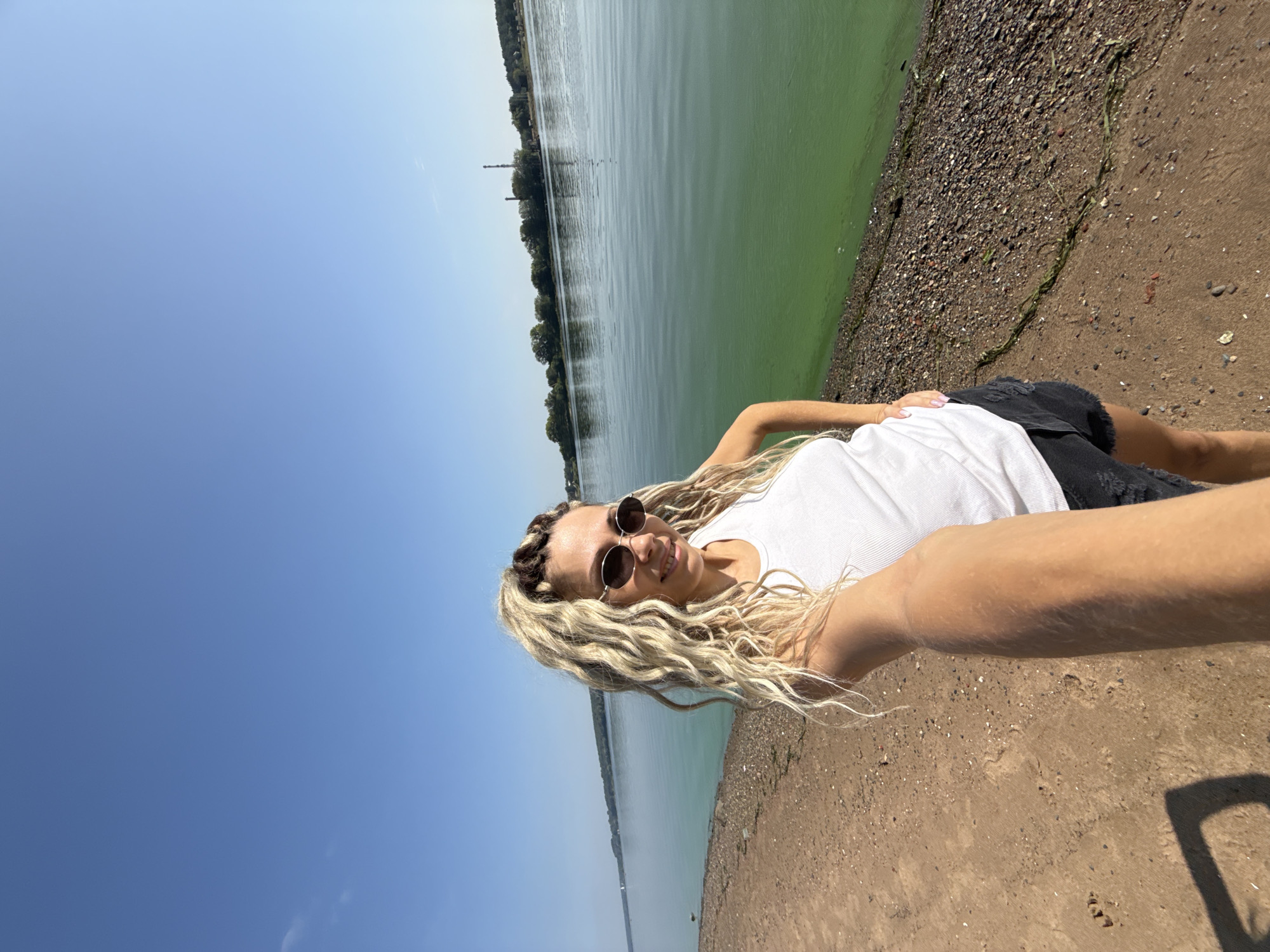 A young woman with blonde dreadlocks and sunglasses is taking a selfie on a sunny day on the shore of a body of water. She is wearing a white tank top and black shorts. The water appears green, possibly due to algae. In the background, there is a line of trees on the opposite shore and a clear blue sky. The location is identified as the city of Rusanovo, Russia. The ground she is standing on is a mixture of sand and small stones.
