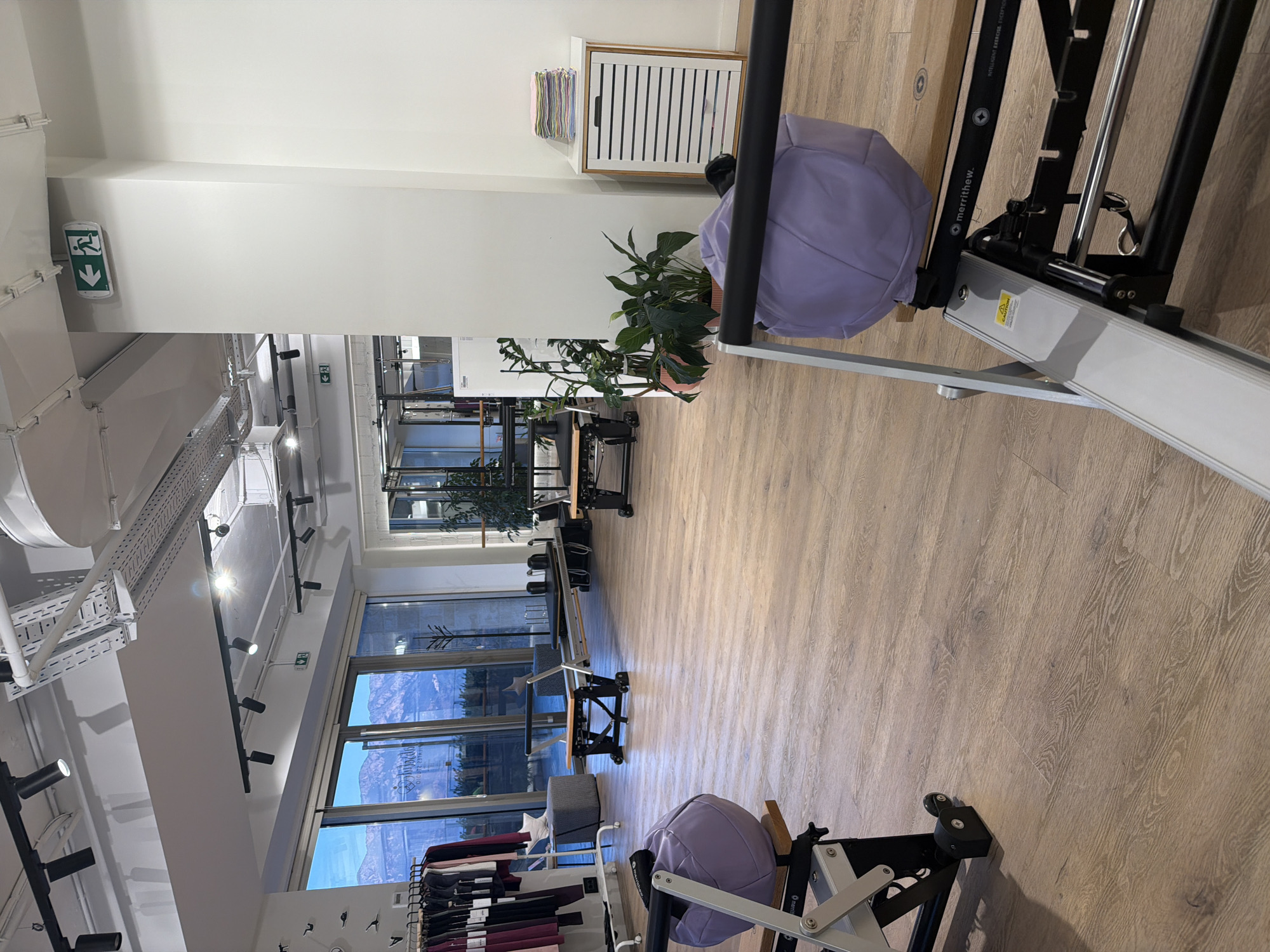 The image depicts the interior of a Pilates studio located in Boreti, Montenegro. The studio features a light wood floor and large windows offering a view of mountains and a body of water, likely the Adriatic Sea.
In the foreground, two Pilates reformers are partially visible. The one on the left has a purple padded piece and silver metal framing. The one on the right is mostly out of frame but shows a black padded surface and a similar metal frame, with the brand name "merrithew." prominently displayed.
Further into the studio, more Pilates reformers are set up, some with wooden platforms and black padded carriages. There is also a rack of dark-colored athletic wear on the left side of the image.
In the mid-ground, a stack of folded towels in various pastel colors is placed on a white cabinet with horizontal black slats. Several potted plants, including a tall Fiddle Leaf Fig, are positioned near the cabinet.
Overhead, track lighting is installed on the ceiling, along with exposed ductwork and conduits, suggesting a modern industrial aesthetic. An illuminated green exit sign with a running figure is visible on the wall. The overall ambiance is clean, spacious, and organized, designed for fitness activities. There are no people visible in the image.
