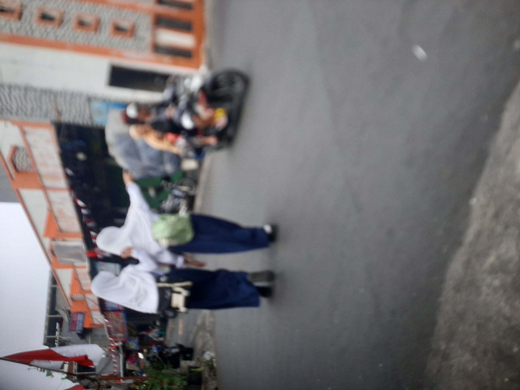 In Central Jakarta, Indonesia, two individuals dressed in white hijabs and dark blue skirts walk on a paved street. The person on the left wears a white hijab and a dark blue skirt, with a white bag slung over their shoulder. The person on the right also wears a white hijab and dark blue skirt, carrying a light green backpack. Behind them, a motorcycle carries two passengers. The buildings lining the street are modern, with orange and white facades. Indonesian flags are visible, suggesting a celebratory or nationalistic context. The scene is slightly blurred, giving a sense of movement and an everyday urban atmosphere.