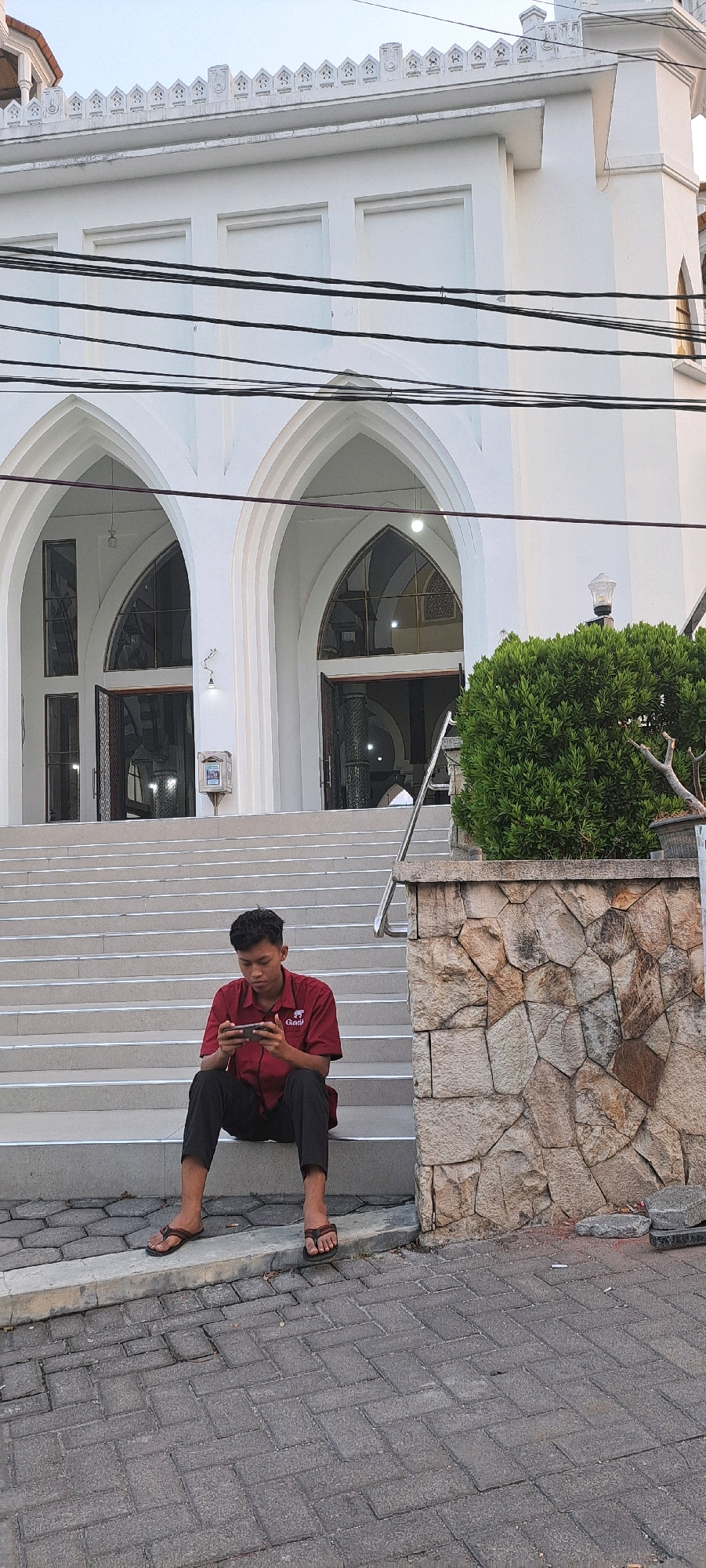 A young man, appearing to be of Indonesian descent, sits on the steps of a white building, possibly a mosque, and plays a mobile game. He is dressed in a red short-sleeved shirt and black pants, wearing brown flip-flops. The building features arched entrances and decorative elements along its roofline. A stone wall with a bushy green plant and a streetlamp stands to the right of the steps. The ground is paved with grey interlocking bricks. The scene is set in Petungasri, Indonesia.