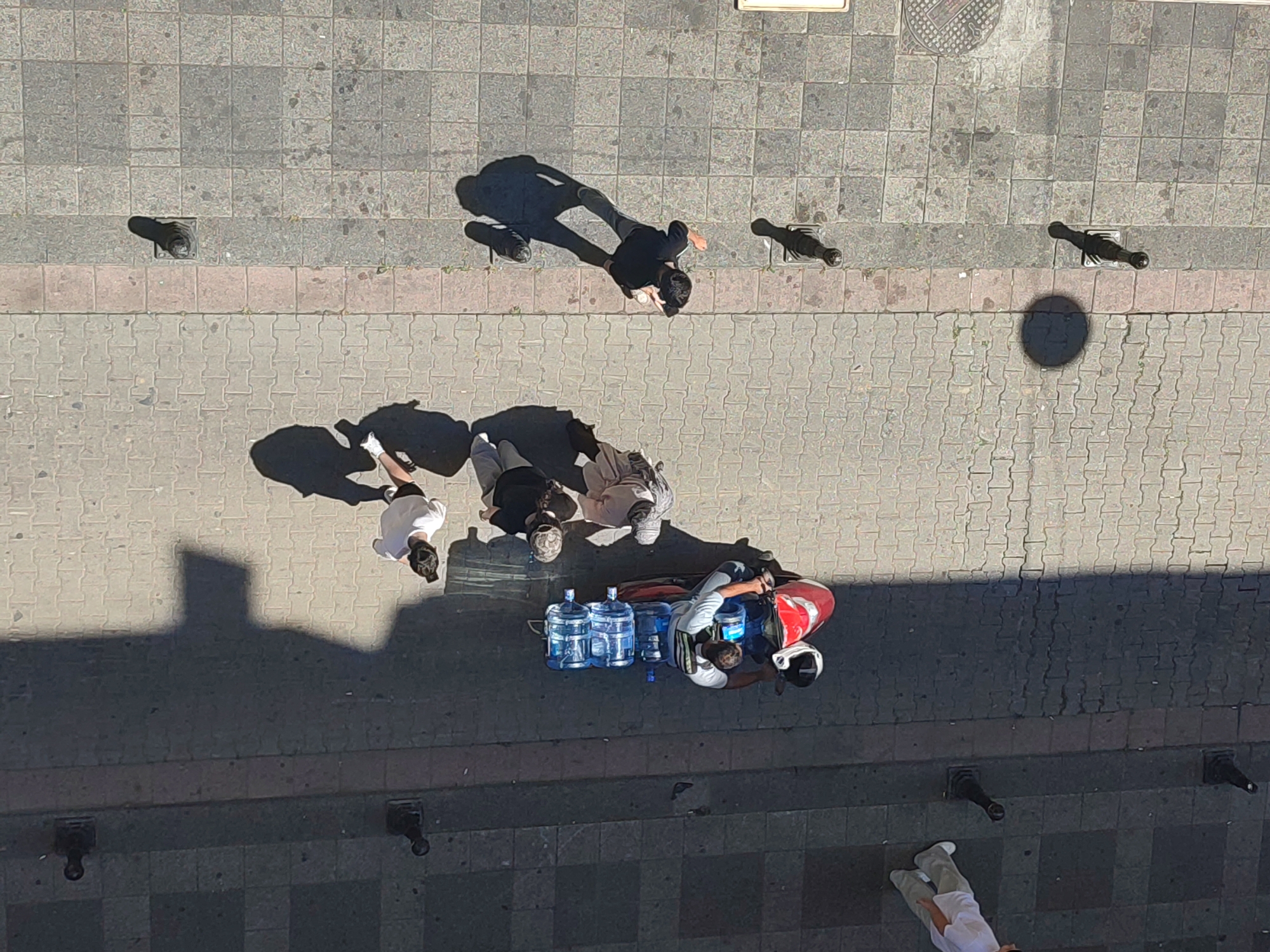 This is an aerial shot looking down at a pedestrian walkway in Kadıköy, Turkey, a busy district on the Asian side of Istanbul. The walkway is paved with interlocking gray and pinkish-brown rectangular tiles. Several people are visible, as are some objects.
In the upper portion of the image, two individuals are walking. One, in a black t-shirt and pants, is casting a long shadow. Nearby, a dark, cylindrical object with a flared top, possibly a bollard or part of a street fixture, is visible. Another similar object is further to the right.
In the middle ground, a group of three people appear to be interacting. One person in a white t-shirt is looking down, while two others are partially obscured.
In the lower middle ground, a person in a white shirt and helmet is next to a red insulated delivery bag strapped to a motorcycle. Several large blue water jugs are arranged in front of the motorcycle. This person is likely a water delivery person.
In the lower part of the image, two more dark, cylindrical street fixtures are visible on the left and right. Further down the walkway, a person wearing light-colored pants and white shoes is walking away from the camera.
The overall scene depicts a typical urban streetscape with people engaged in everyday activities. The lighting suggests it is daytime, with strong shadows indicating a relatively high sun angle.