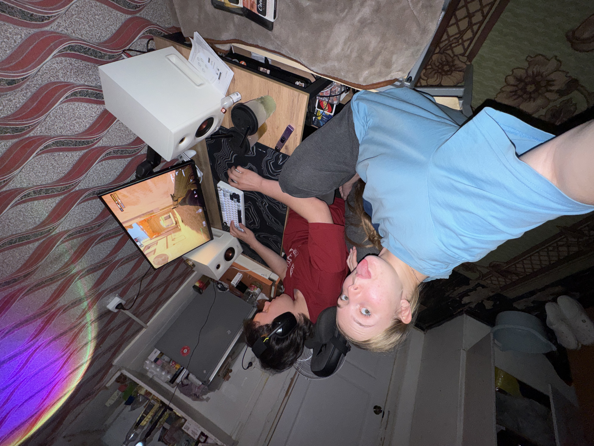 Two people are in a room, rotated in the frame so that the camera is oriented with what would normally be to the left. A woman in a blue shirt is visible in the top right corner sticking out her tongue and facing the camera. Beneath her, a man in a red shirt wears headphones and has his hands on a keyboard. They are positioned near a computer gaming setup. A white projector sits atop a desk, casting light onto the wall, and a computer monitor displays a video game. A desk and shelves are visible in the background and to the left. Various items, including containers and decorative objects, are scattered throughout the space. The walls are decorated with wavy red patterns. A rug with floral designs is partially visible at the top-right. The room appears to be in a domestic setting.
No location is mentioned or implied.