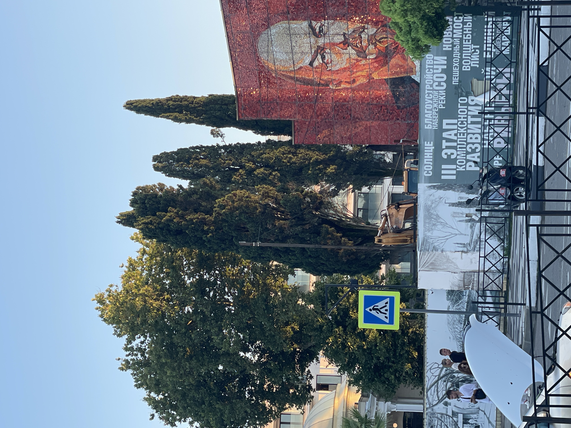 The image shows a street scene in Sochi, Russia, dominated by a large mosaic mural of Vladimir Lenin on the side of a red brick building. In front of the building, a billboard displays text in Russian, including "SOLNTSE" (Sun), "BLAGOUSTROYSTVO NABEREZHNOY REKI SOCHI" (Embankment Development of the Sochi River), and "PESHEKHODNYY MOST VOLSHEBNYY LIST" (Pedestrian Bridge Magic Leaf).
Several tall cypress trees are positioned in front of the mural, obscuring parts of the building. To the left of the cypress trees, a large leafy tree with scattered sunlight on its foliage stands tall. Behind the trees, the upper floors of modern buildings with balconies are visible.
In the foreground, a white car with its hood open is parked at a crosswalk, indicated by a pedestrian crossing sign with a blue background and a white pictogram of a person walking. A black metal fence runs along the sidewalk.
Several people are gathered near the car and the billboard. Some individuals are walking, while others appear to be observing the scene. A yellow construction vehicle is partially visible behind the billboard. The sky is clear and blue, suggesting it is daytime.