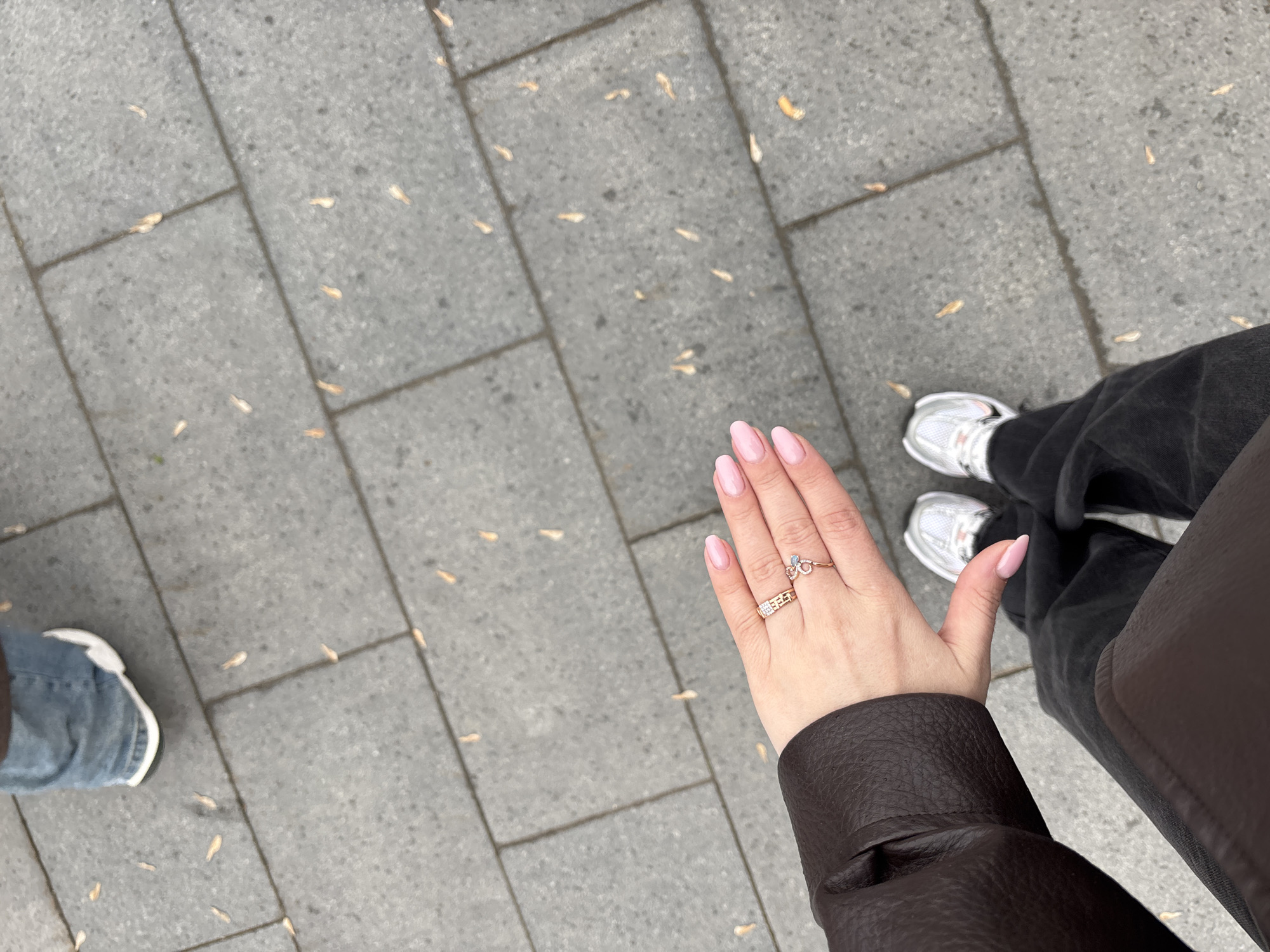 The image shows a person standing on a gray brick sidewalk with scattered leaves. The person is wearing a brown leather jacket, black jeans, and white sneakers. The person's hand is visible, adorned with two gold rings and light pink nail polish. Another person's foot, clad in a blue denim jean and white sneaker, is visible in the lower left of the frame. The location is Moscow, Russia.