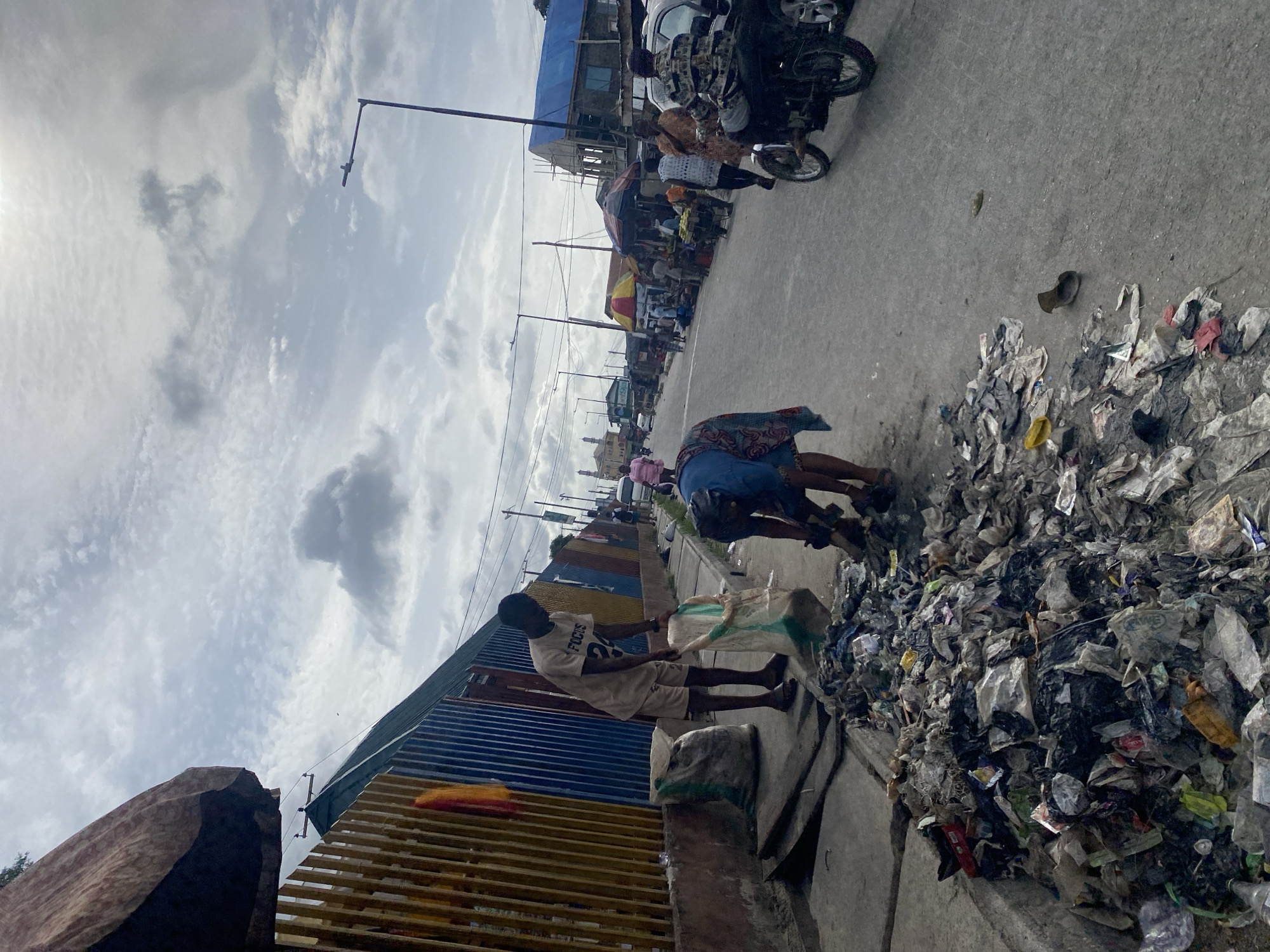 In Ado, Nigeria, the image depicts a street scene with two people scavenging through a large pile of mixed waste, including plastic bags and discarded items, on the side of a paved road. A young man in a light-colored shirt and shorts is holding a plastic bag, seemingly collecting items from the waste. An adult, wearing blue and patterned clothing, is also sorting through the trash. In the background, a street market scene unfolds, with people, motorcycles, and structures visible along the road. Streetlights are visible in the distance, and a cloudy sky is overhead.