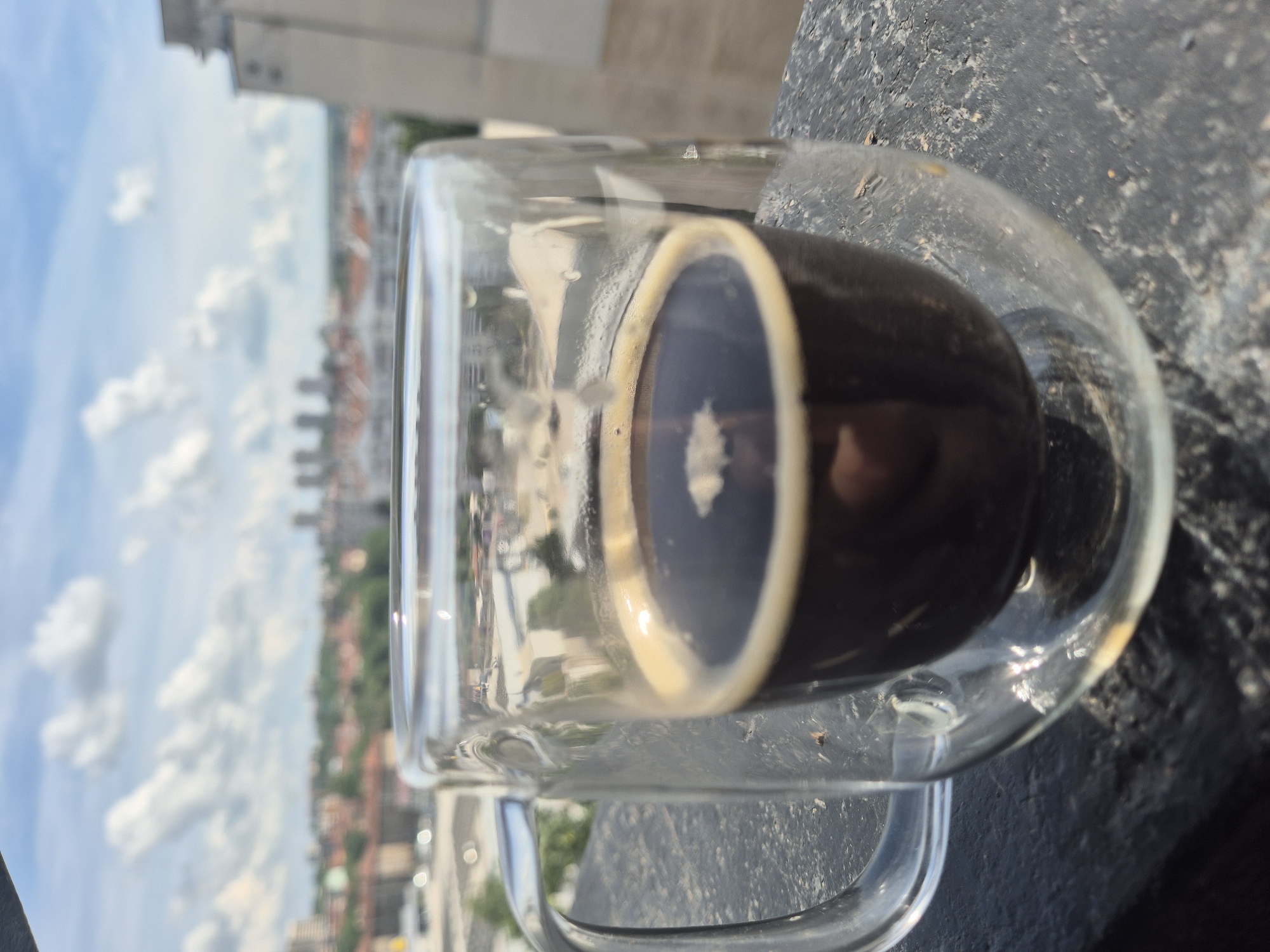 A double-walled glass mug filled with dark coffee sits on a textured dark gray surface. The mug has a clear handle and a light-colored foam on top of the coffee. In the background, a cityscape is visible under a bright blue sky with scattered white clouds. Buildings of various heights and architectural styles, some with orange rooftops, form the urban landscape. Trees are interspersed throughout the city. The lighting suggests it is daytime.