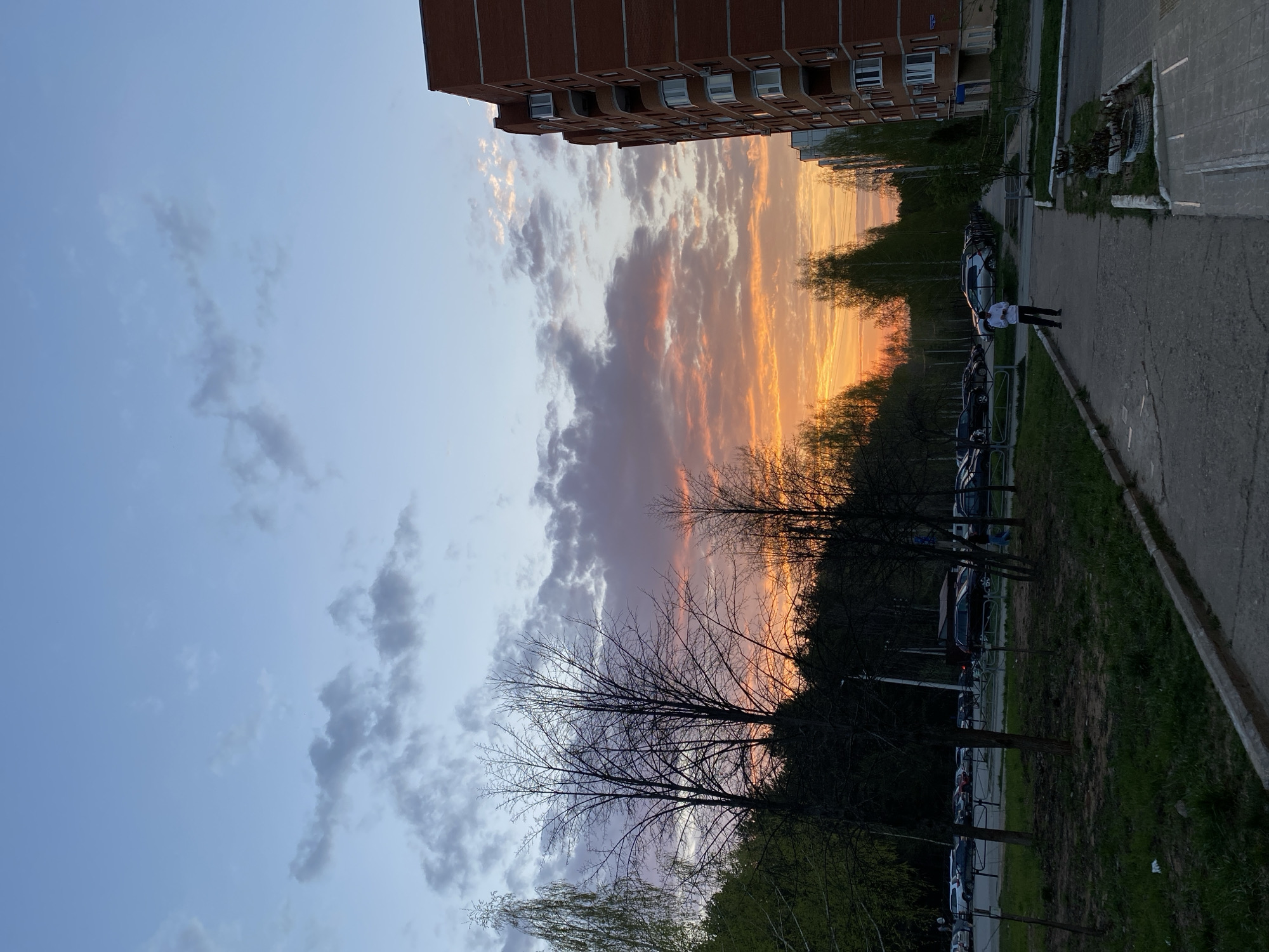 Here is a description of the image:
The image is taken in what appears to be Chaikovsky, Russia. It shows an urban environment at sunset. On the right is a sidewalk and parking lot adjacent to a grassy area with trees. Several cars are parked in the lot. Two people are seen walking on the sidewalk, near the top right of the frame. A large brick apartment building is in the background. The sky is divided between a blue sky with grey clouds and a vibrant orange sunset obscured partially by clouds. The trees in the middle ground are silhouetted against the colorful sky.