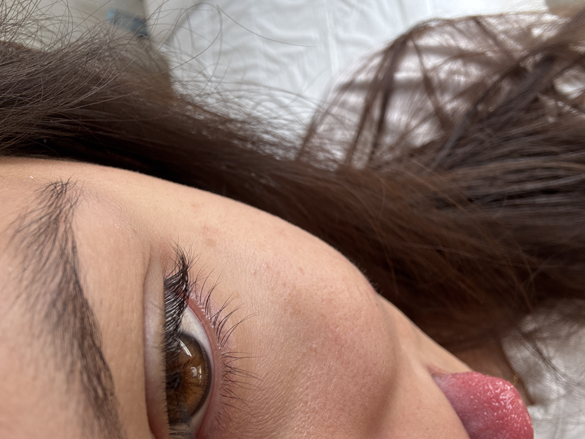 A close-up shot depicts a person lying on a white surface, possibly a bed. The individual has dark brown hair and a prominent right eye with brown irises. Visible facial features include an eyebrow and a portion of the tongue, which is pink and textured. The setting appears to be indoors, with no discernible details linking it to San Francisco Ocotlán, Mexico.