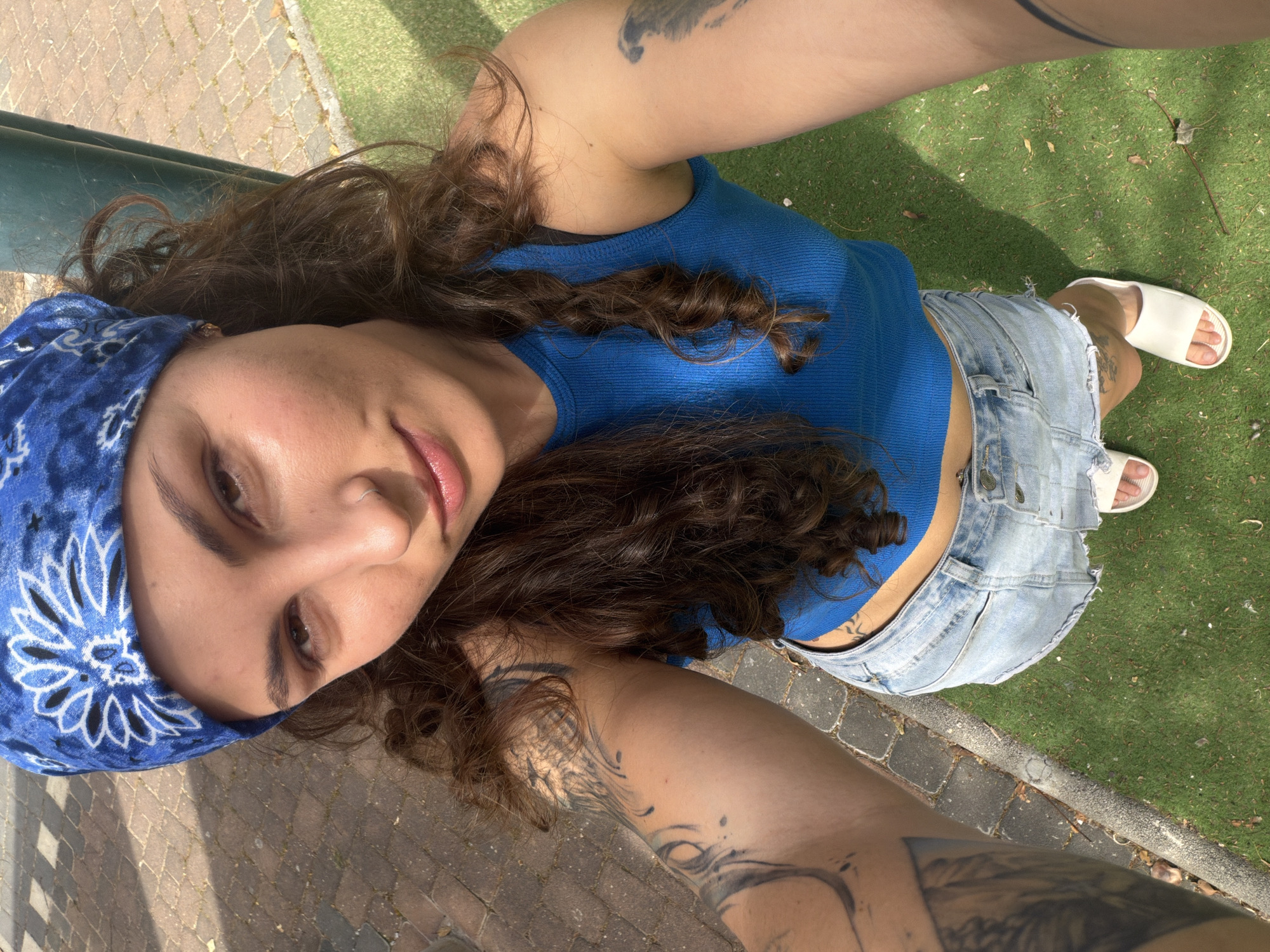 A young person, likely female, is captured in a high-angle, low-perspective selfie, seemingly reclining or lying down outdoors in Kiryat Motzkin, Israel. She looks directly at the camera with a relaxed expression and a slight smile.
She has long, dark, curly hair that flows around her head and shoulders. Her attire includes a blue bandana with a white floral or paisley pattern, a ribbed blue tank top, and light-wash, distressed denim shorts. She wears white slide sandals on her feet. A silver nose ring is visible, and multiple tattoos are prominent on her arms, shoulders, and potentially her lower back or side.
The background shows a paved or brick pathway on the left and artificial green turf or grass on the right. A dark green metal rail or bench structure runs horizontally in the upper left. The scene is brightly lit by natural sunlight, indicating it is daytime, with shadows cast clearly on the ground. There is no visible text in the image.