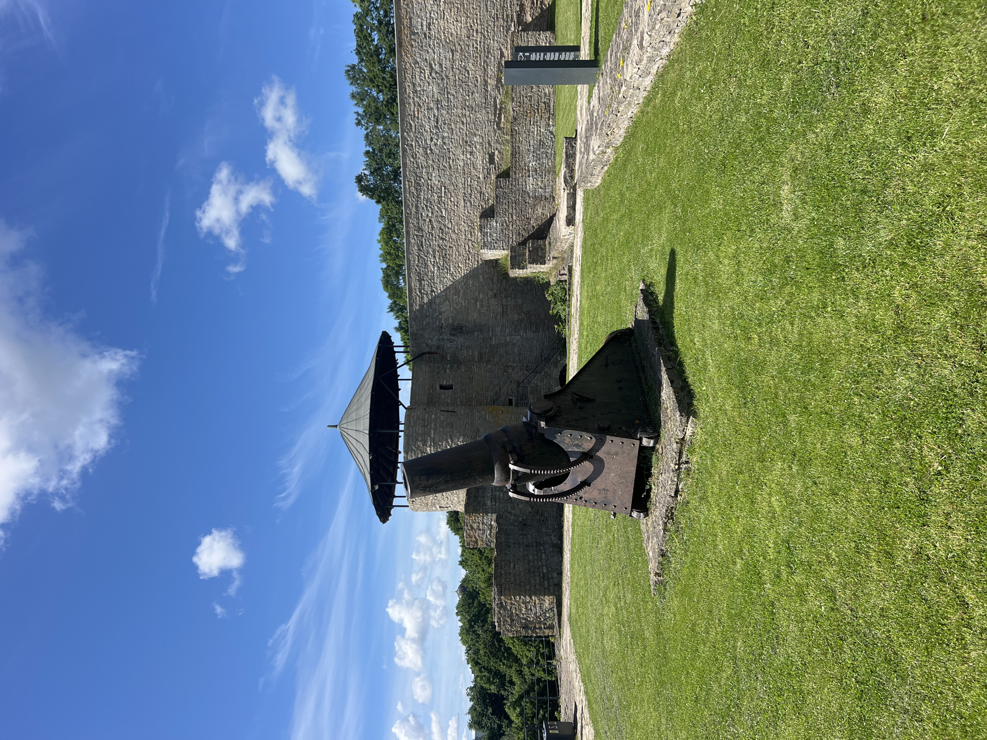 This image, taken in Narva, Estonia, depicts a historical outdoor scene on a sunny day. The primary subjects are a weathered, dark-colored cannon situated on a grassy area, and the stone walls of what appears to be a fortification or castle. The cannon is positioned on a metal base with visible gears and is pointed towards the left of the frame. Above and behind the cannon, a conical, dark-colored roof structure is visible, supported by wooden beams.
The background is dominated by the ancient, rough-hewn stone walls of a building, suggesting a historical site. Trees with lush green foliage are visible beyond the walls, and a bright blue sky with scattered white clouds fills the upper portion of the image. The grass in the foreground is vibrantly green and well-maintained, casting shadows from the cannon and the walls.
The overall atmosphere is tranquil and evokes a sense of historical significance. The time of day appears to be mid-morning or early afternoon, indicated by the clear sky and the angle of the shadows. No people are visible in the frame. There is no discernible text in the image.