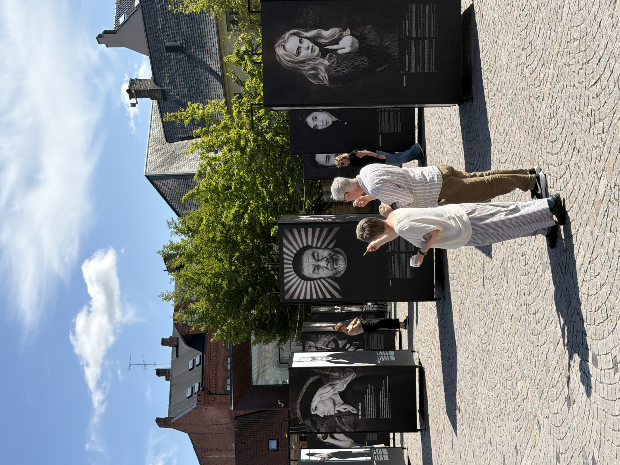 An outdoor photography exhibition is taking place on a cobblestone plaza in the city of Lund, Sweden. The exhibition features large, black-and-white portraits displayed on tall, freestanding panels. The portraits are of various individuals, including a woman wearing a hat and the singer Zara Larsson giving the middle finger. Several people are viewing the exhibition. In the foreground, a person with short hair and wearing a white shirt and striped pants is looking at a portrait and holding a white cup. To their right, a man wearing a striped shirt and brown pants is standing and looking at the same portrait, holding a small object in his hand. In the background, buildings with red brick and dark roofs are visible, along with lush green trees. The sky is blue with some white clouds. The overall atmosphere is casual and public, suggesting a cultural event or art display.