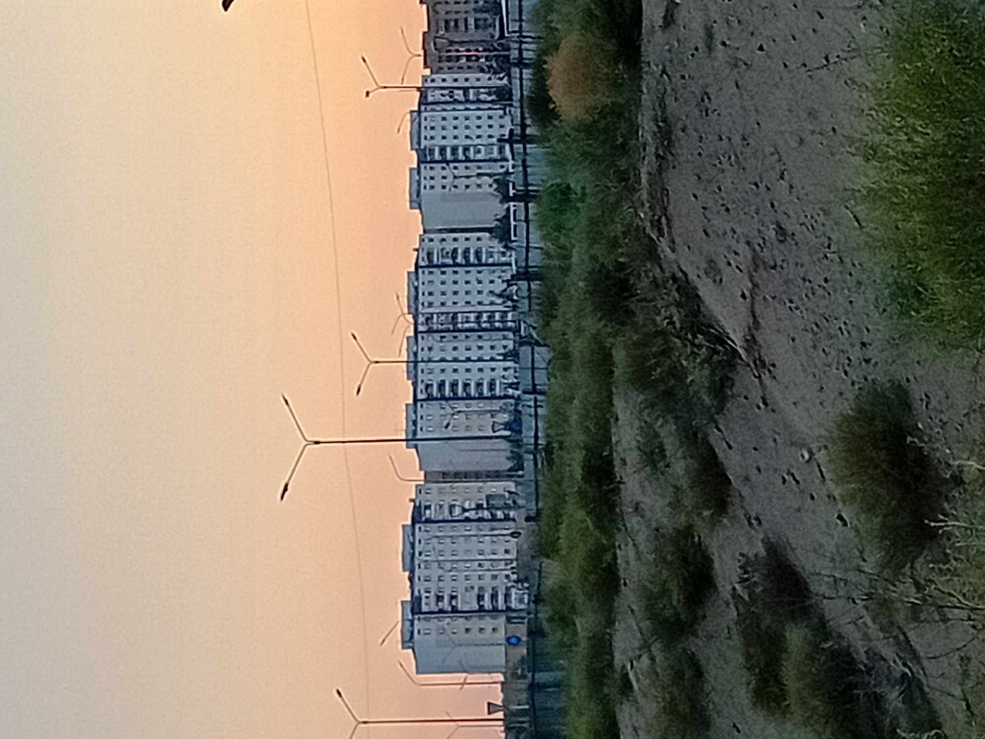 The image depicts a row of identical, multi-story apartment buildings under a hazy, pale orange sky, likely during either sunrise or sunset. The buildings are light-colored, with numerous windows arranged in a grid pattern. They appear to be of Soviet-era architecture. Several streetlights with Y-shaped fixtures are visible in front of the buildings, interspersed with some trees. In the foreground, a barren, sandy slope is covered with sparse, dry vegetation and some low-lying green bushes. The overall impression is one of a residential area on the outskirts of a city. This scene is characteristic of the residential neighborhoods in Semey, Kazakhstan, particularly the areas where such uniform apartment blocks are common.