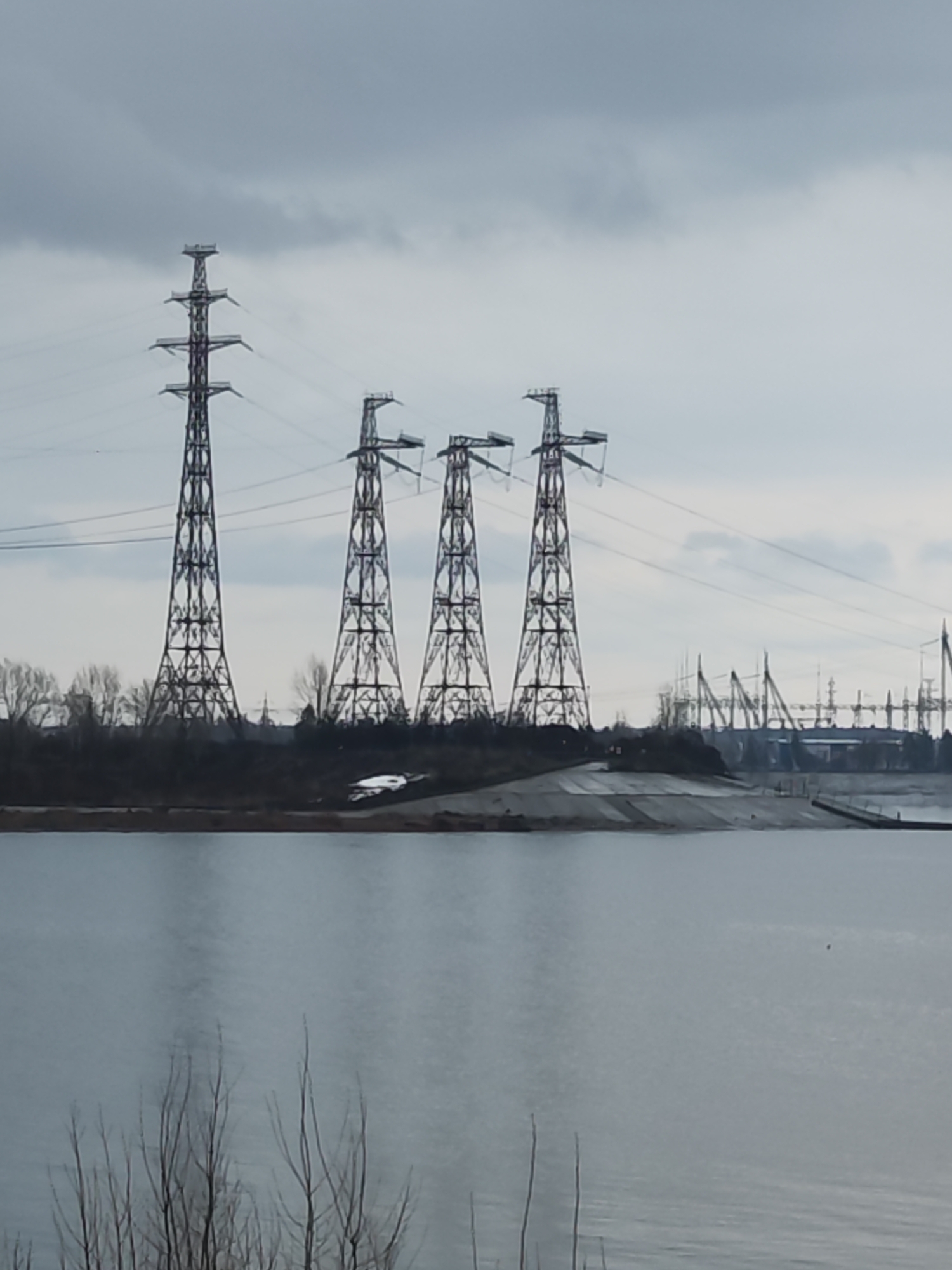 The image shows a landscape in Chaikovsky, Russia. Four electrical transmission towers are the prominent features in the midground. The towers are black metal lattice structures with interconnected cables running between them. In the background, additional power structures and industrial elements are visible. The foreground consists of a body of water, with barren trees in the lower portion of the frame. A shoreline is visible between the water and the towers, consisting of a concrete bank. The sky is overcast, suggesting a cloudy day.
