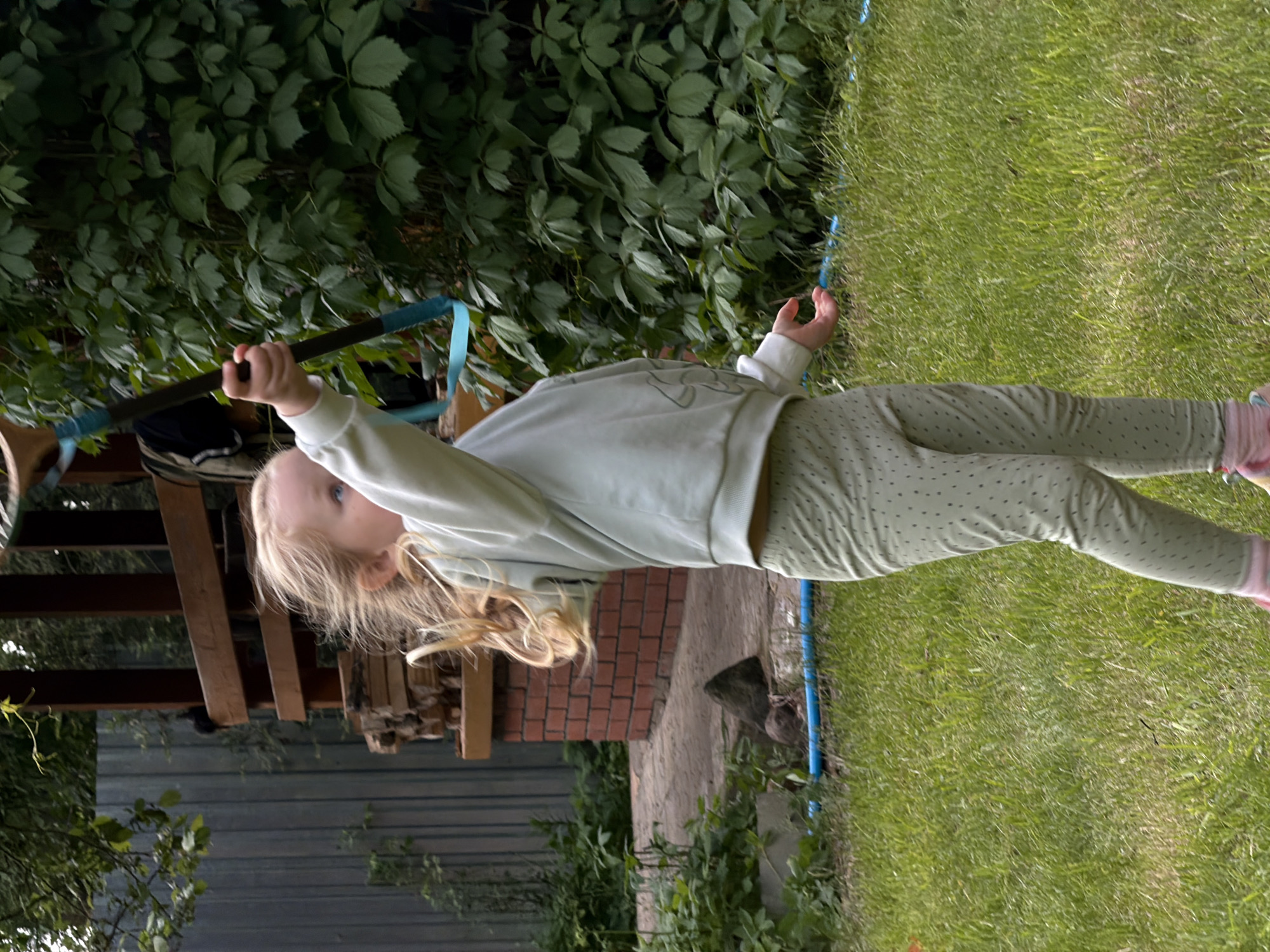 A young Caucasian girl with blonde hair is swinging on a makeshift swing constructed from a dark-colored pole with blue straps, seemingly in a garden or yard. The girl is wearing a light-colored long-sleeved top and green pants with dark dots. She is holding onto the pole with one hand and extending the other. The background includes a dense growth of green foliage, a wooden structure, a brick wall, and a section of a grey fence. The location is presumed to be in Udelnaya, Russia.
