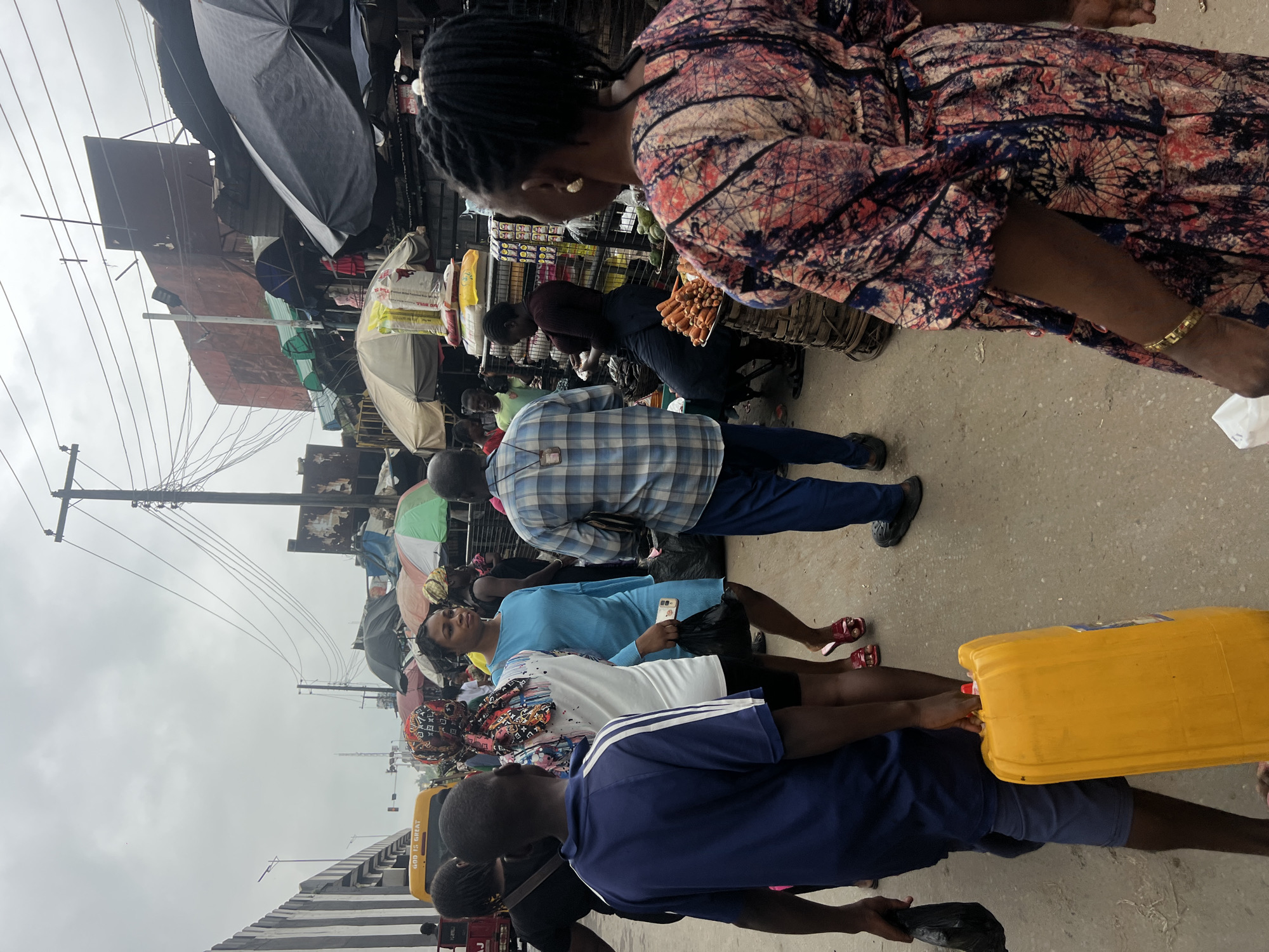 The image depicts a busy outdoor market scene in Nkpor, Nigeria, under an overcast sky. In the foreground, a young boy wearing a navy blue t-shirt with white stripes walks away from the camera, carrying a large yellow jerrycan. To his left, a person in a black t-shirt and dark shorts is also walking away, holding a black plastic bag. In the center of the image, a woman in a bright blue dress walks towards the right, holding a black plastic bag and a smartphone. Behind her, a man in a blue and white plaid shirt is walking to the left. Further back, other individuals are visible navigating the crowded market, with stalls and various goods displayed. The market environment includes overhead wires, umbrellas providing shade, and structures made of corrugated metal. A yellow truck with the words "GOD IS GREAT" is partially visible on the left side of the image. The ground is dirt and appears to be a well-trafficked area.