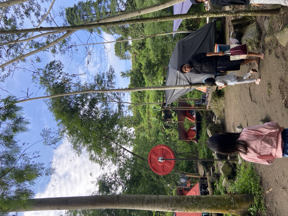 A person with a child stands outside a black tent near a path. Another person, wearing a pink shirt, has their back to the camera. A red weight hangs from a metal bar between the trees. The background consists of a dense green forest and a blue sky with white clouds visible through the treetops. Tall, slender trees line the perimeter.
