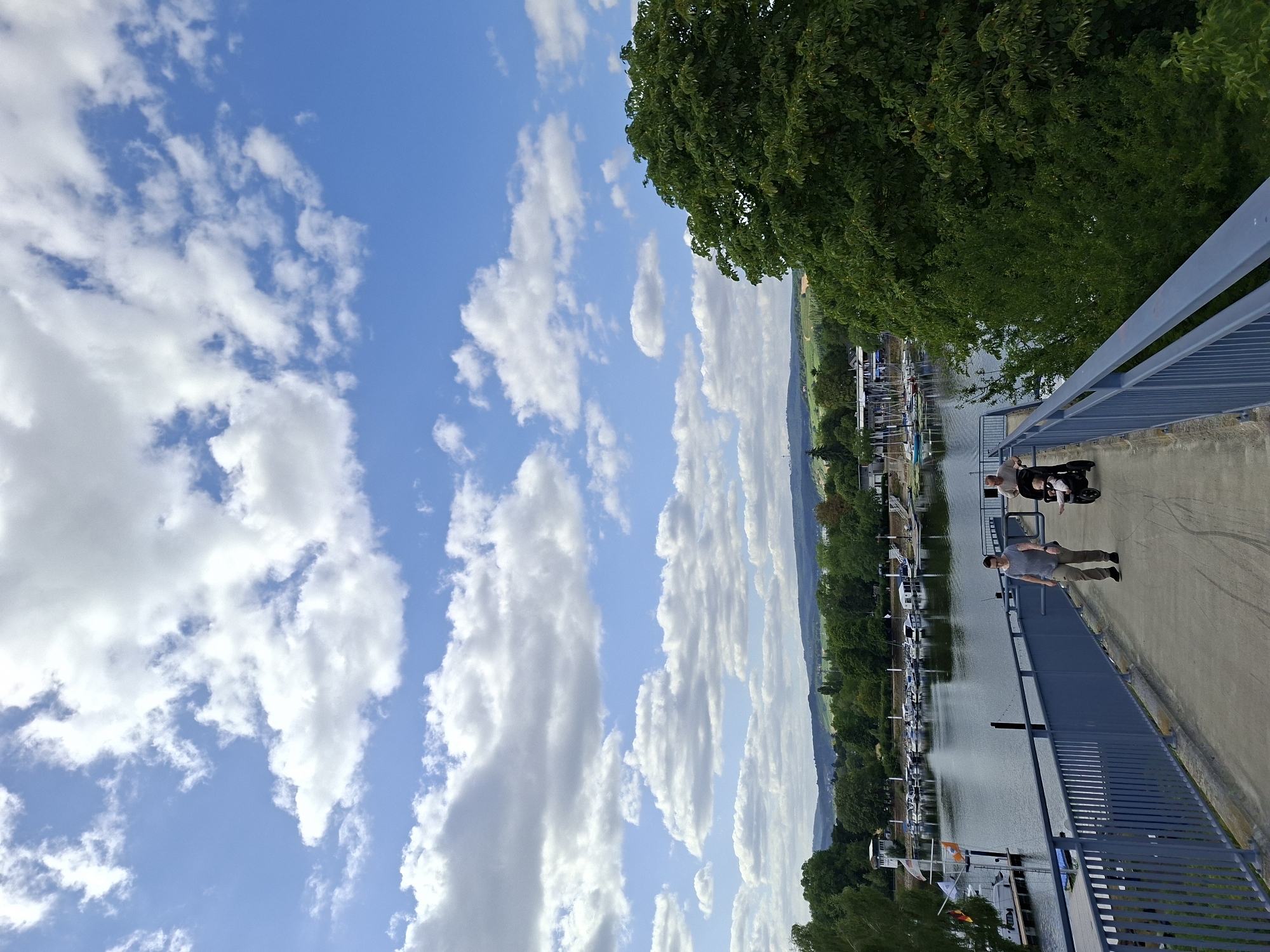 A high-angle, full shot captures a bright, partly cloudy day over a waterfront scene. The foreground features a blue metal railing and a concrete pathway, with two adults and a baby in a stroller walking away from the viewer. One adult, wearing a gray t-shirt and khaki pants, walks slightly ahead, while the other, in a light brown t-shirt and dark pants, pushes the stroller. Behind them, the pathway curves, offering a view of a calm body of water lined with docked boats and a marina. Lush green trees and rolling hills form the middle and background, with a glimpse of buildings nestled amongst the foliage. The sky is a vibrant blue, dotted with fluffy white clouds. This scene depicts the area near the Wiesbaden Biebrich yacht club.
