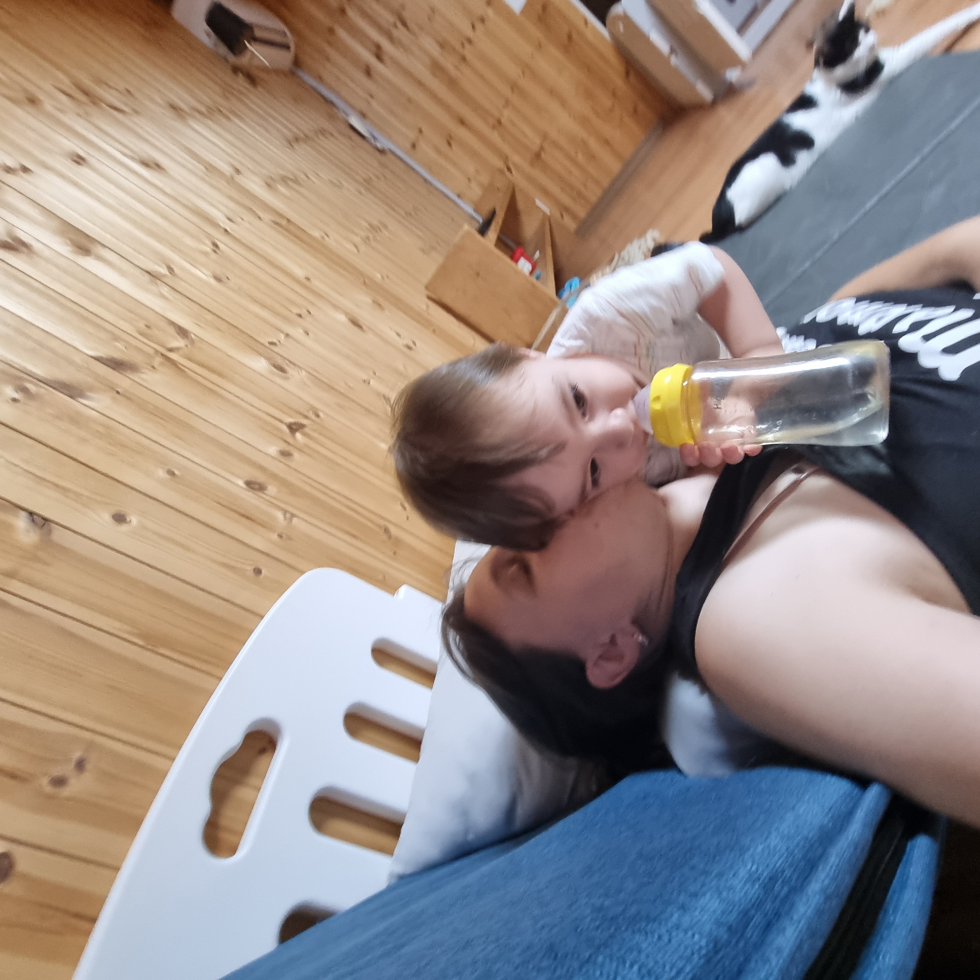 In a room with wood-paneled walls, a woman is seen kissing a baby on the cheek. The baby is holding a bottle with a yellow nipple. The woman is wearing a black tank top, and the baby is wearing a white shirt. A blue blanket is visible on the left side of the image, and a white crib is partially visible in the background. A black and white cat is lying down in the lower right portion of the frame. The environment appears to be a domestic setting, possibly a bedroom or living area. The image does not provide any specific information about the location being Dunnae-myeon, South Korea.