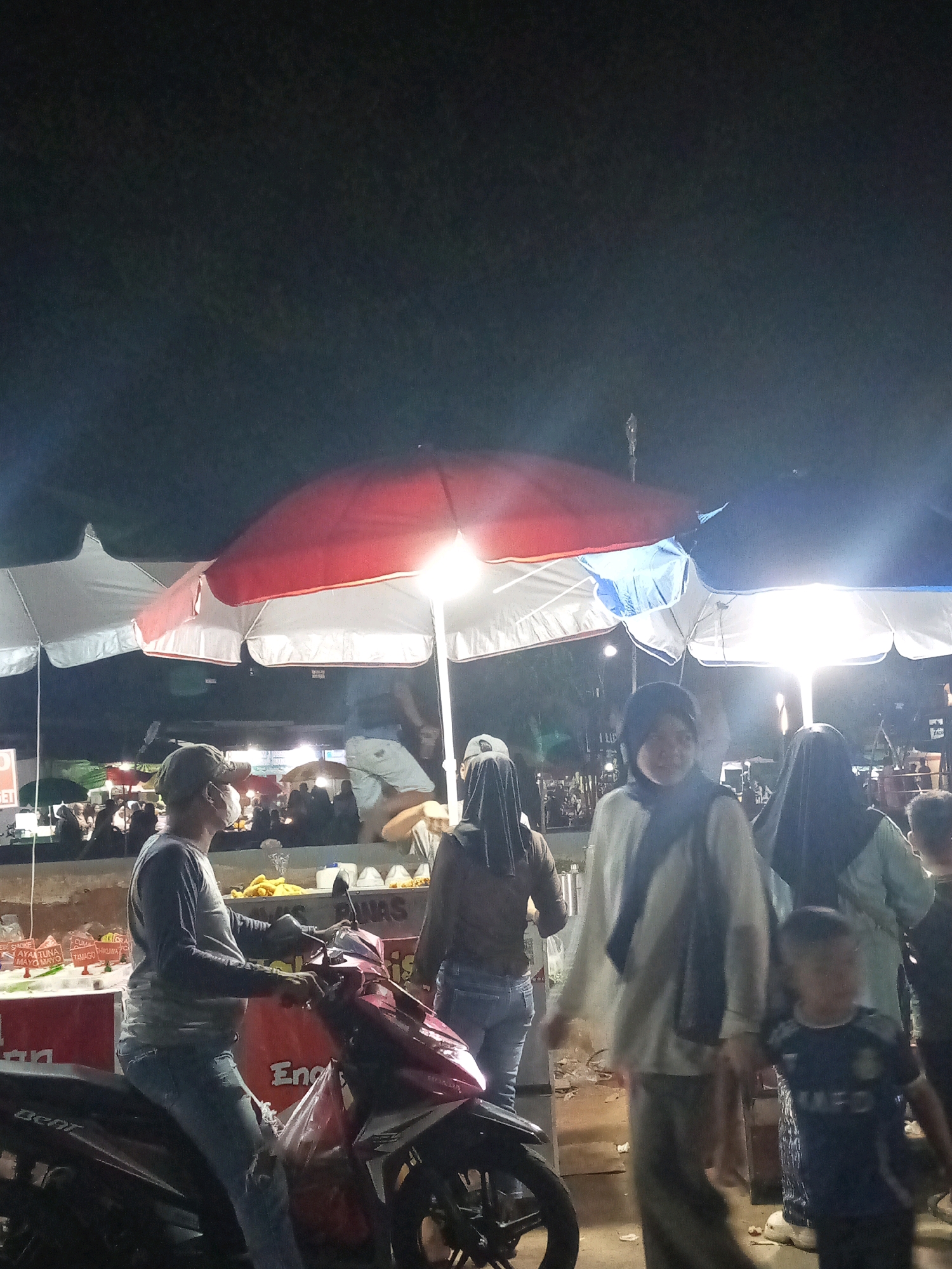 This is a nighttime photograph of an outdoor market, likely a food bazaar, in Bekasi, Indonesia. The scene is illuminated by bright lights, including those from the market stalls and overhead lamps. In the foreground, a man is seated on a red motorcycle, wearing a long-sleeved shirt, jeans, and a cap. He is partially obscured by the motorcycle and appears to be observing the market.
Behind him, several market stalls are visible, covered by large red and white umbrellas. These stalls appear to be selling food, with containers of ingredients or prepared dishes displayed. Several people are browsing the stalls.
Further into the background, more market activity is evident, with crowds of people and various stalls. The overall atmosphere suggests a bustling evening market. The image captures a slice of local life and commerce in Bekasi.