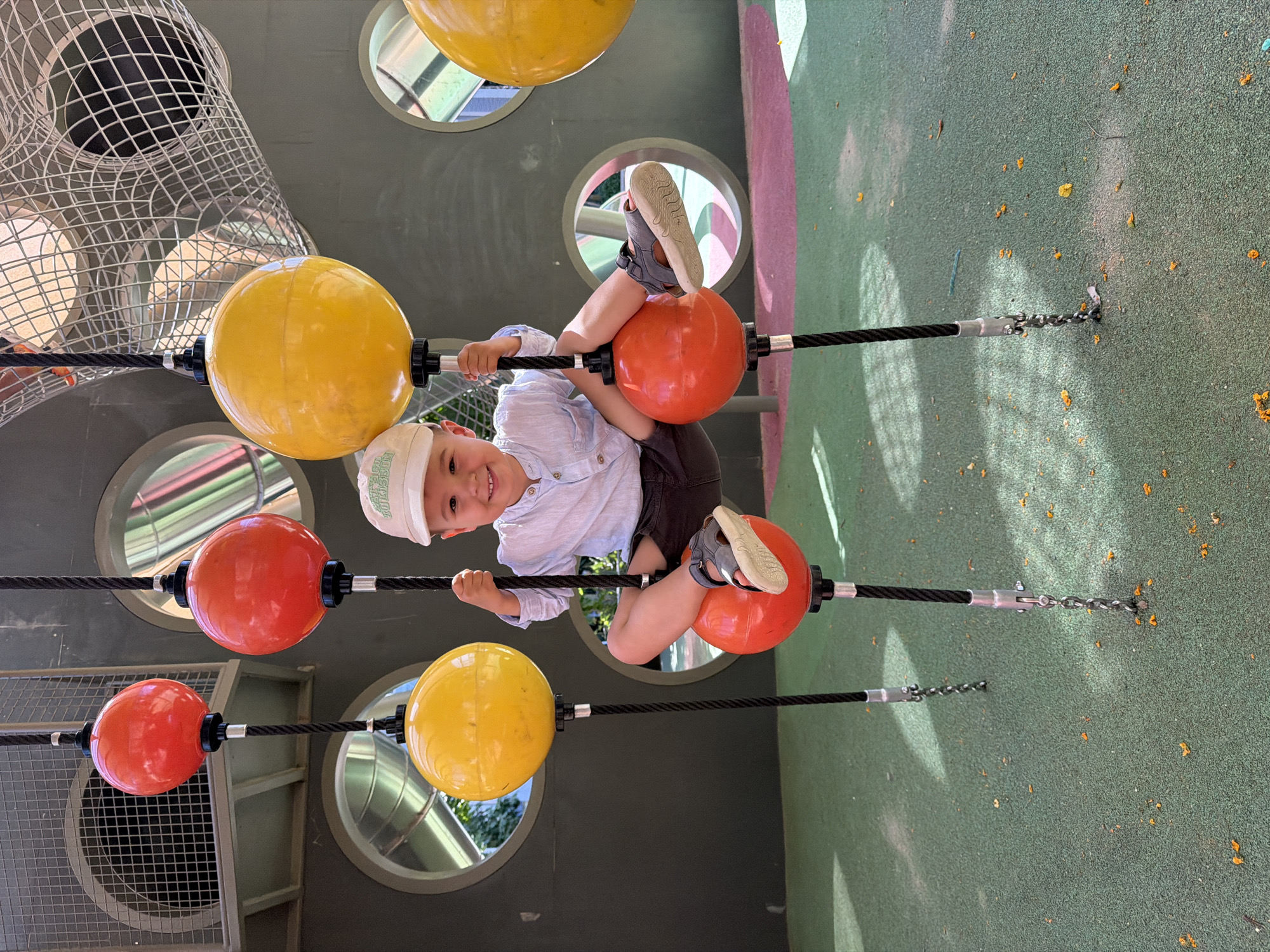 A young boy, appearing to be around three years old, is playfully sitting on a piece of playground equipment. He is wearing a light-colored collared shirt, dark shorts, a light-colored baseball cap, and dark sandals. The boy is straddling a black rope with red spherical objects attached to it, with one leg resting on a red sphere and the other extended slightly. He is smiling at the camera. The background features a dark-colored wall with circular openings and a net-like structure above. The ground beneath the playground equipment is a green rubberized surface with some scattered yellow and orange leaf-like debris. The scene suggests an outdoor playground environment. The image was taken in Barcelona, Spain.
