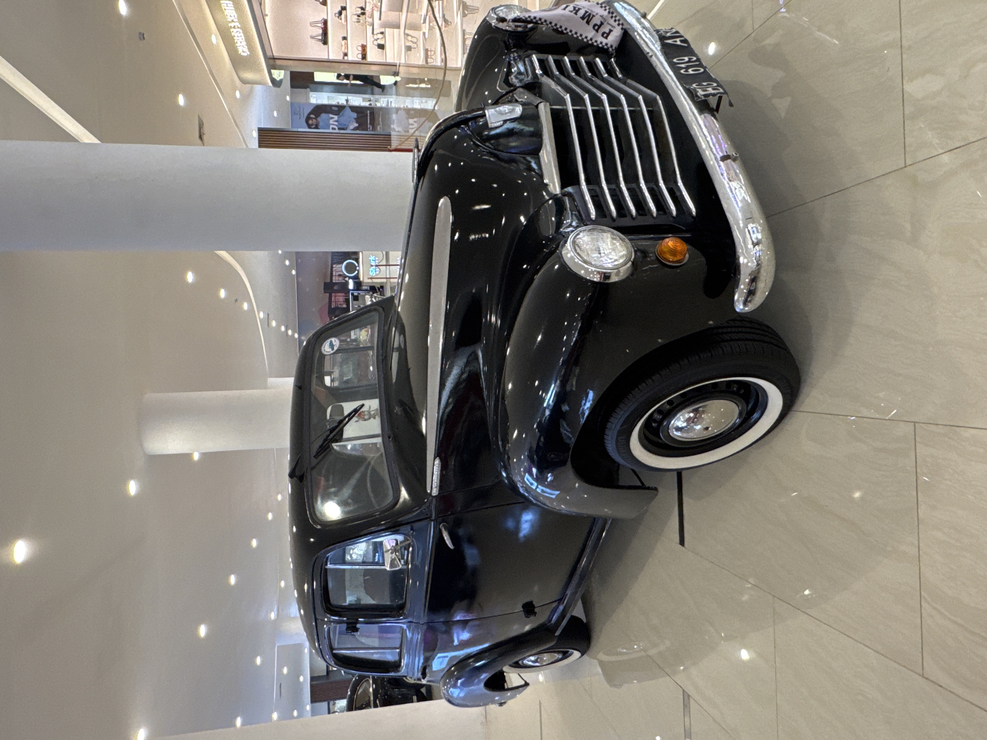 The image depicts a vintage black car, likely a classic Austin taxi, prominently displayed indoors on a light-colored tile floor. The car is positioned at an angle, showcasing its front grill, headlights, and part of the side. The license plate is partially visible. In the background, a shopping mall environment is discernible, with shops and interior lighting. The car is indoors, likely on display. The location is Kuta, Indonesia.