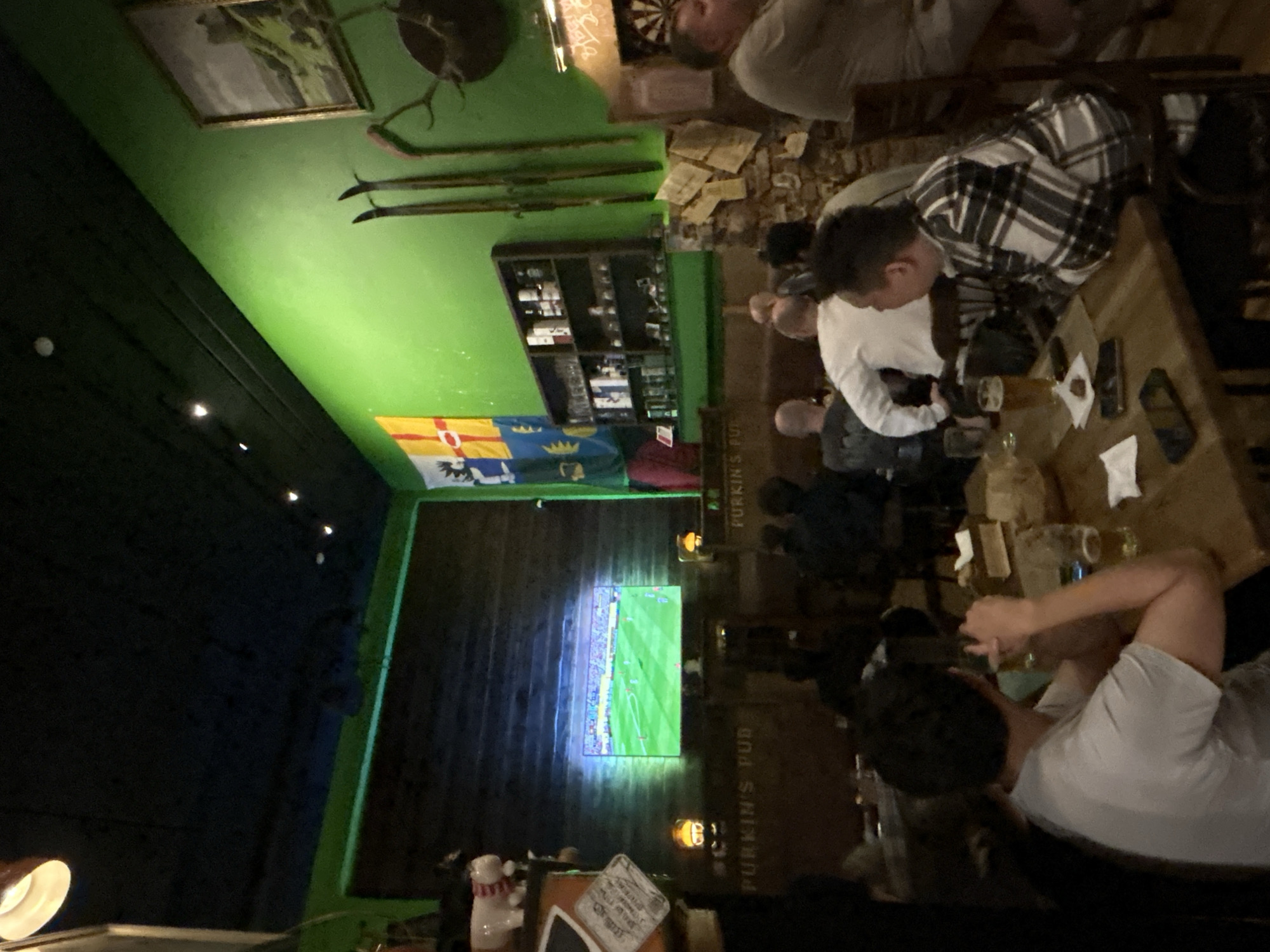 The image depicts the interior of a dimly lit pub named "PURKIN'S PUB". The walls are painted a vibrant green, and the ceiling is black. A large television screen on the far wall displays a soccer match. Several patrons are seated at tables, watching the game and socializing. Some are drinking beer. The decor includes flags, mounted antlers, and shelves stocked with bottles. The overall atmosphere appears casual and relaxed, typical of a sports bar. There is no indication of this location being in Moscow, Russia.