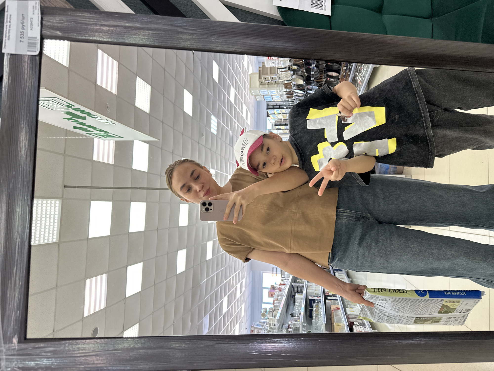 The image captures a mirrored scene of a woman and a child in a store, presumably in Chaikovsky, Russia. The woman, holding a smartphone, is taking a selfie with the child who is giving the peace sign. The reflection shows store shelves, indicating a shopping environment, and a price tag on the frame suggests they are examining a mirror for sale. The woman wears a tan top and blue jeans, while the child is dressed in a dark shirt with a yellow design and a white hat with a pink brim. The store's ceiling has a pattern of square panels, and the environment is well-lit. A newspaper is also visible in the reflection.