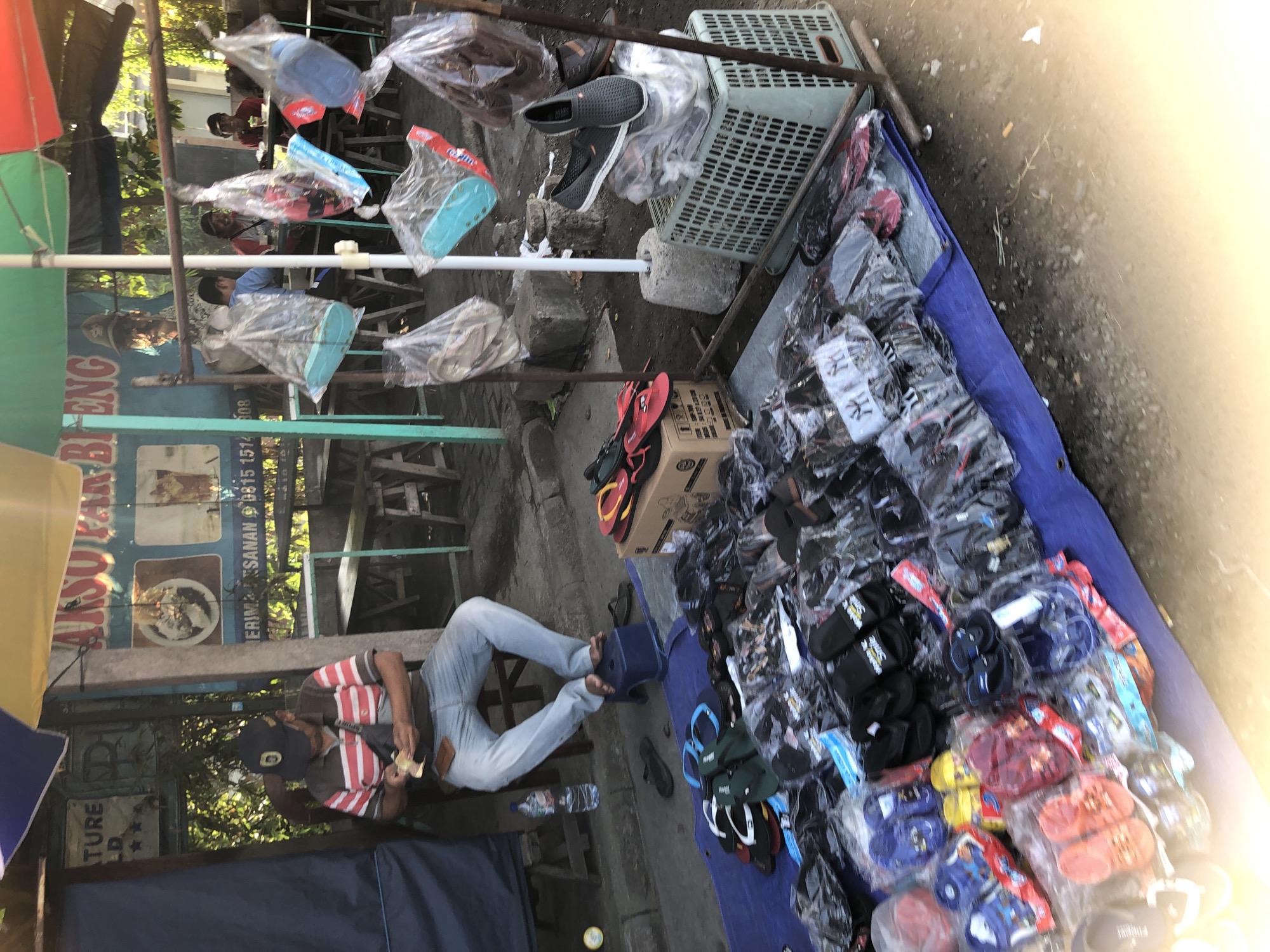 The image depicts an outdoor market stall in Balongdowo, Indonesia, likely selling footwear. In the foreground, a blue tarp is spread on the ground, laden with various pairs of flip-flops and sandals, many packaged in clear plastic. A man wearing a striped polo shirt, jeans, and a black baseball cap is seated on a stool, holding money in his hands. Behind him, a food stall with a banner reading "AKSO BAR BENG" is visible, along with seating areas and other patrons. Several plastic bags containing footwear are hanging from a metal frame, and a grey plastic basket filled with shoes sits on the ground. The scene appears to be on a paved surface, with some debris scattered around. Sunlight is casting strong shadows, indicating it is daytime.