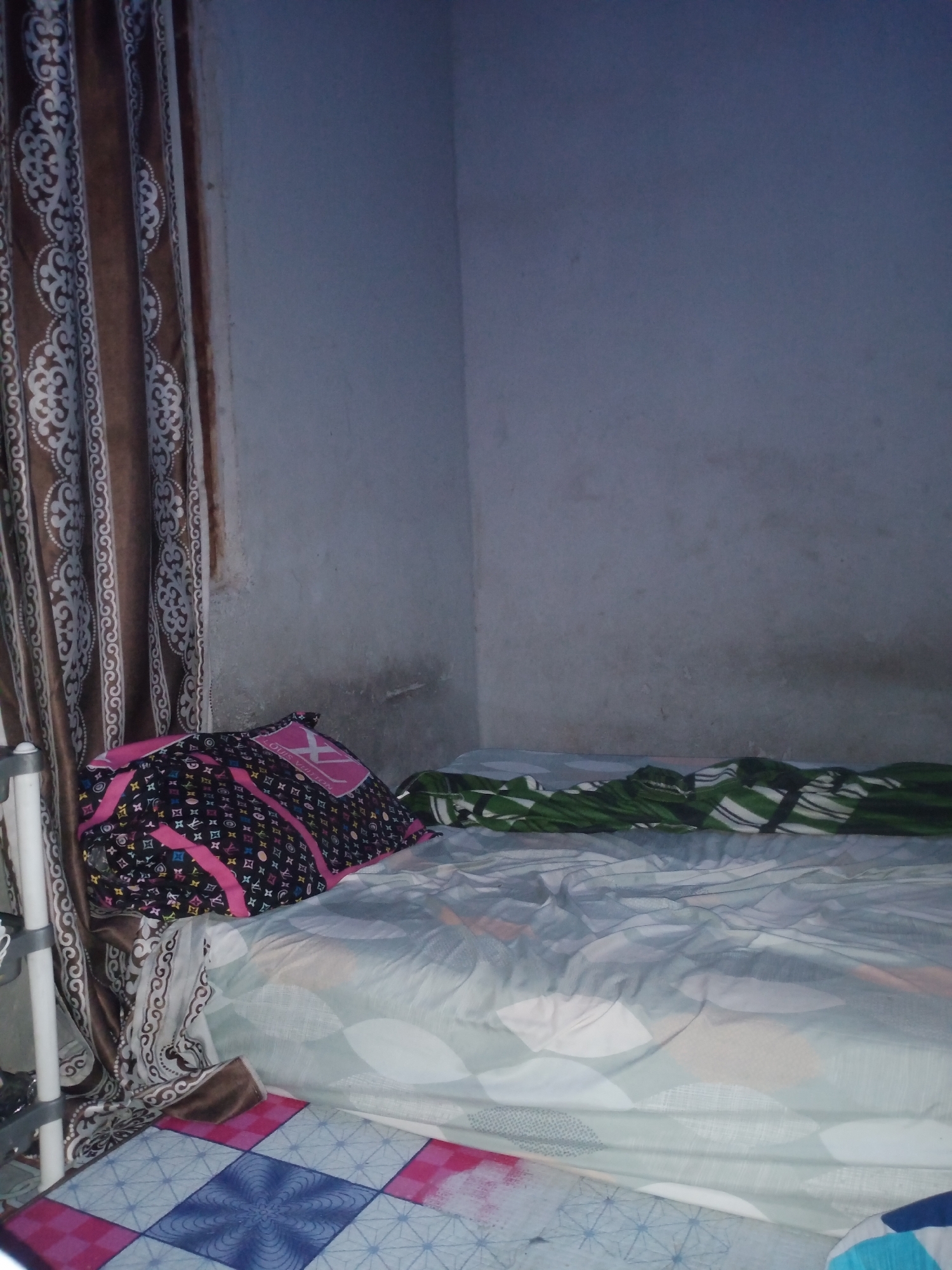The image shows a bed in a dimly lit room, likely a bedroom. The bed has several patterned covers and pillows. On the left side of the bed, a patterned curtain is visible. The walls appear to be a light gray color and show some signs of wear or dirt. The overall impression is that of a simple, possibly unmade bed in a residential setting. The scene is consistent with a bedroom in a dwelling in Jalingo, Nigeria, given the mention of the city.