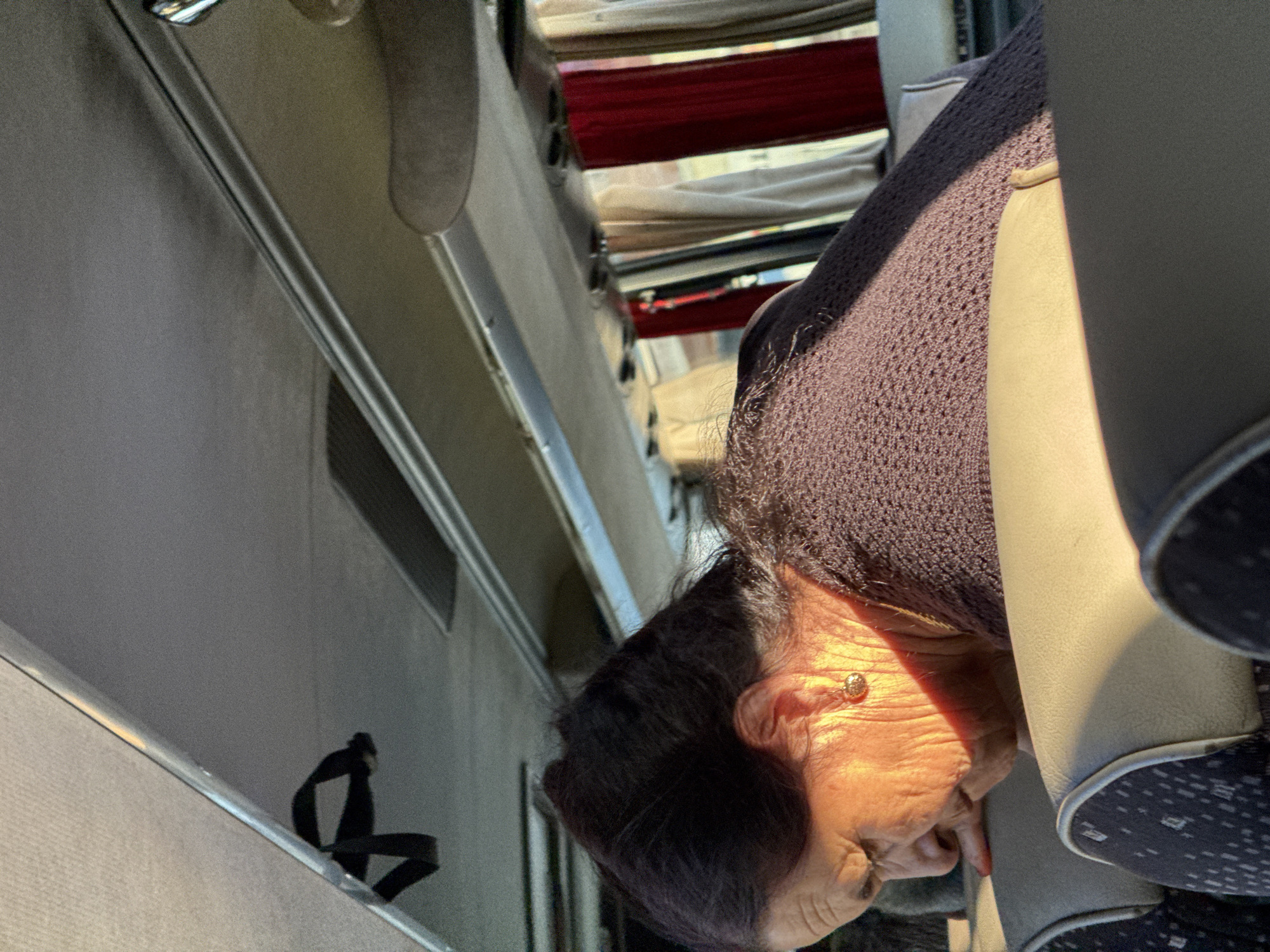 The image shows an elderly woman with dark hair, wearing a dark knitted sweater and a gold earring. She is seated on a bus, with her head tilted down and her hand resting on the back of a seat. Sunlight is illuminating the side of her face, highlighting the wrinkles on her skin. The interior of the bus is visible, with beige seats, grey overhead compartments, and red and beige curtains covering the windows. The lighting suggests it is daytime, possibly during sunrise or sunset. There is a black strap hanging from the ceiling of the bus. The image does not contain any information that would suggest a specific location in Omurtag, Bulgaria.