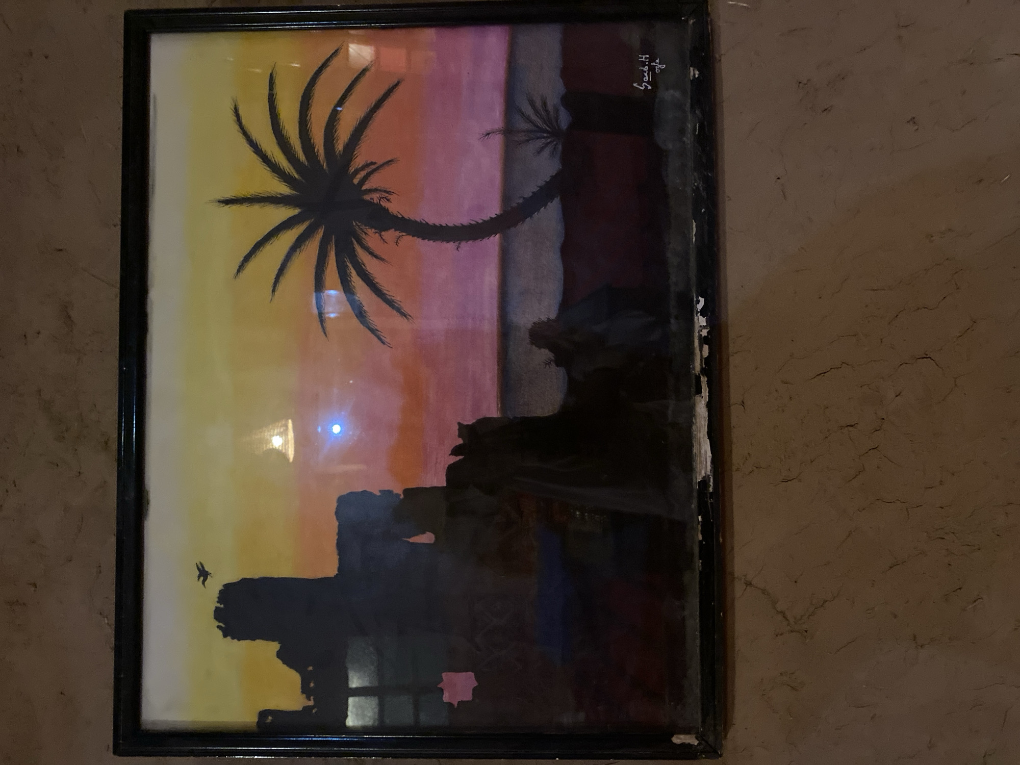 This is a framed painting of a desert sunset scene, likely depicting a location in Agafay, Morocco. The painting features a silhouette of ancient ruins on the left, with a bird in flight against a gradient sky of yellow, orange, and pink. A large palm tree dominates the right side of the composition, with its fronds spread wide. In the foreground, silhouetted figures are visible, suggesting human presence. A smaller palm tree is also depicted on the right. The painting is signed in the lower right corner. The artwork is displayed on a textured, earthy-toned wall.