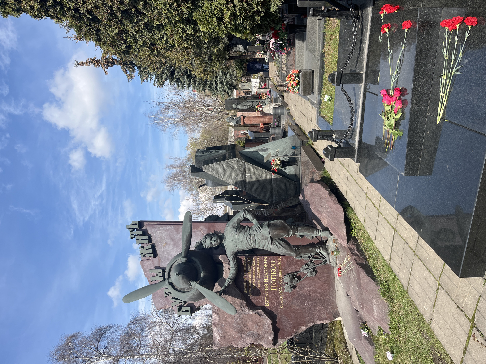 The image shows a monument at the Novodevichy Cemetery in Moscow, Russia. The monument is dedicated to Vitaly Ivanovich Popkov, a decorated Soviet military aviator and twice Hero of the Soviet Union. The monument features a bronze statue of Popkov, an airplane propeller, and a stone backdrop. Other graves and tombstones are visible in the background. Red flowers are arranged on the top of one of the graves. The sky is blue with white clouds.