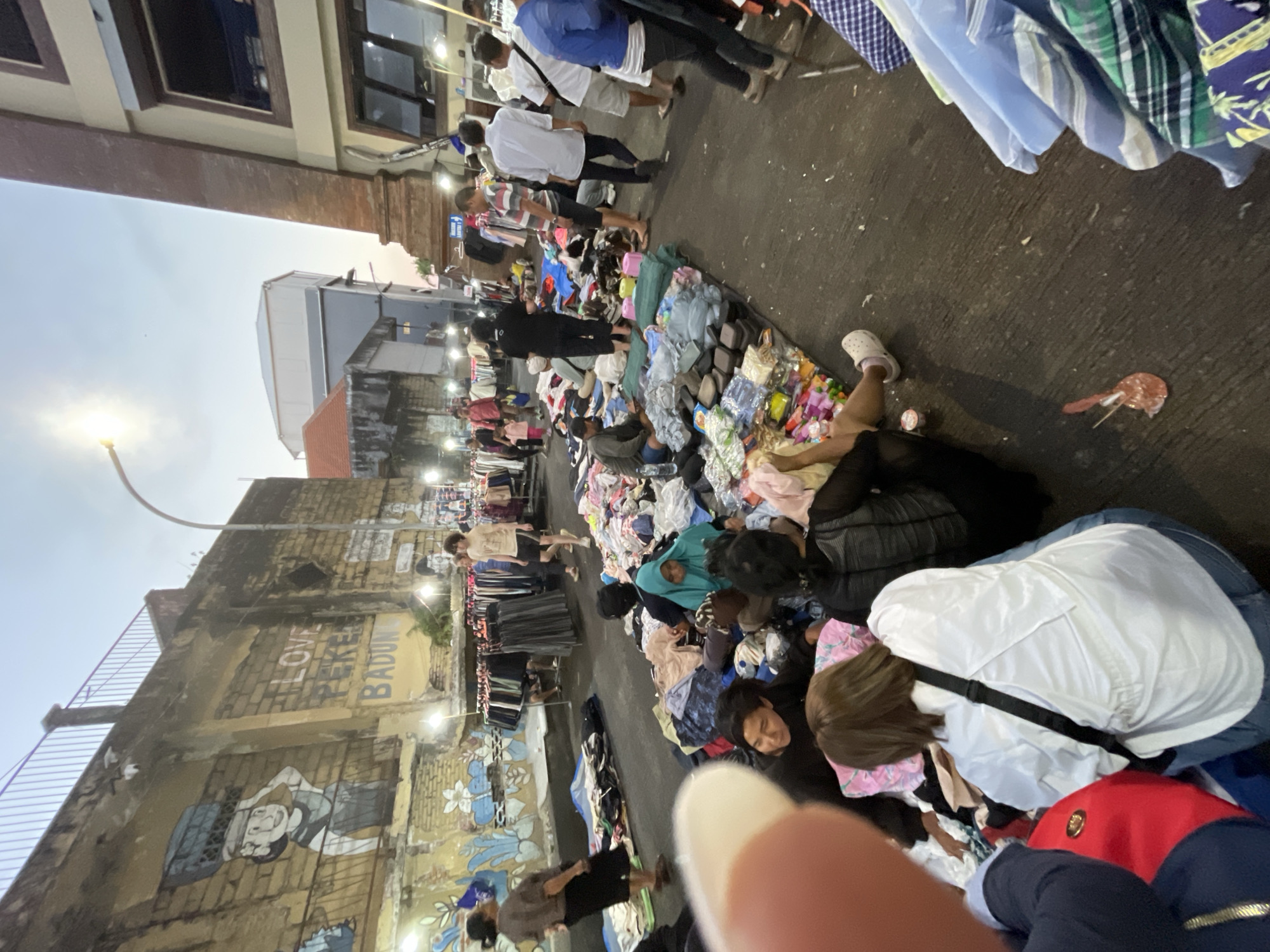 The image captures a bustling street scene in Denpasar, Indonesia, likely at dusk, as indicated by the soft, warm lighting and the sky's gradient. The location appears to be an open-air market, possibly a night market or a roadside stall area, given the rows of clothing racks and the spread of goods on the ground. A prominent feature is a weathered brick wall adorned with vibrant graffiti, including a depiction of a person carrying a load on their head and the words "LOVE PEKE BADUNG."
Numerous people are engaged in various activities associated with a market. Some are browsing through piles of clothing laid out on tarps on the asphalt road, while others stand or sit amongst the merchandise. Several individuals are actively selling or arranging items. The clothing displayed includes a variety of garments, some neatly hung on racks and others piled high on the ground, suggesting a focus on apparel.
The environment is characterized by the presence of streetlights illuminating the area, and in the background, various buildings of different architectural styles are visible, some appearing to be shops or commercial establishments. The overall impression is one of lively commerce and community interaction within a dynamic urban setting.